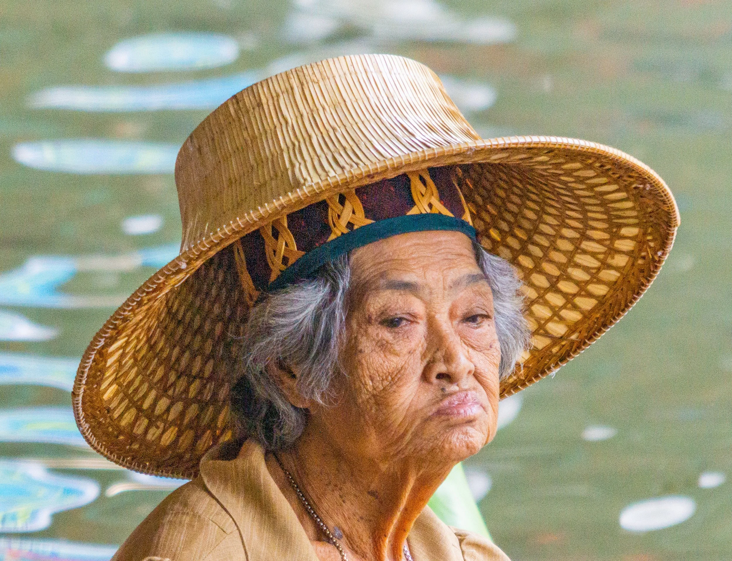 Portrait of an elderly woman wearing a woven hat beside a canal in Thailand.