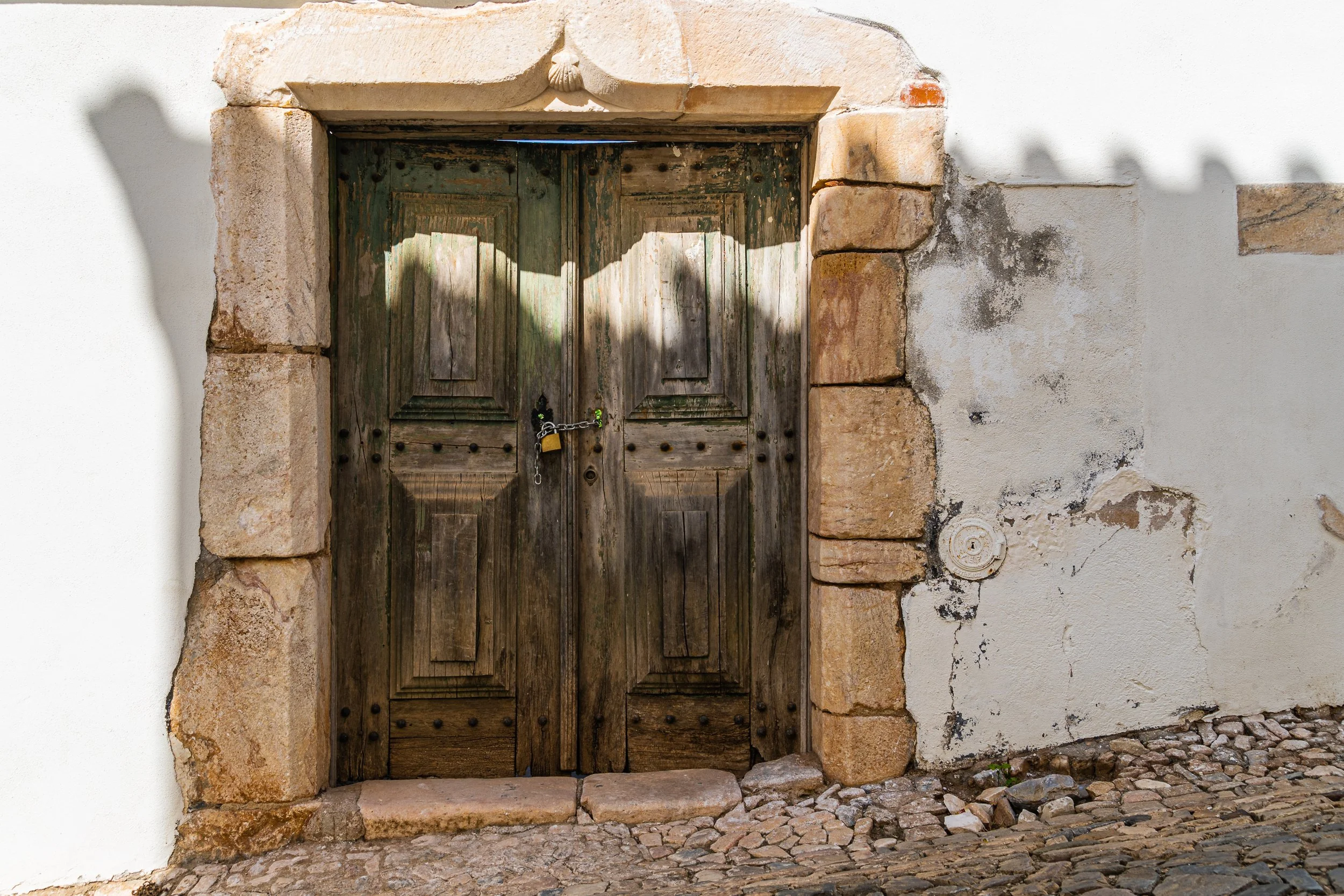 Weathered wooden double doors framed by carved stone, secured with a small padlock along a cobblestone street in Portugal.