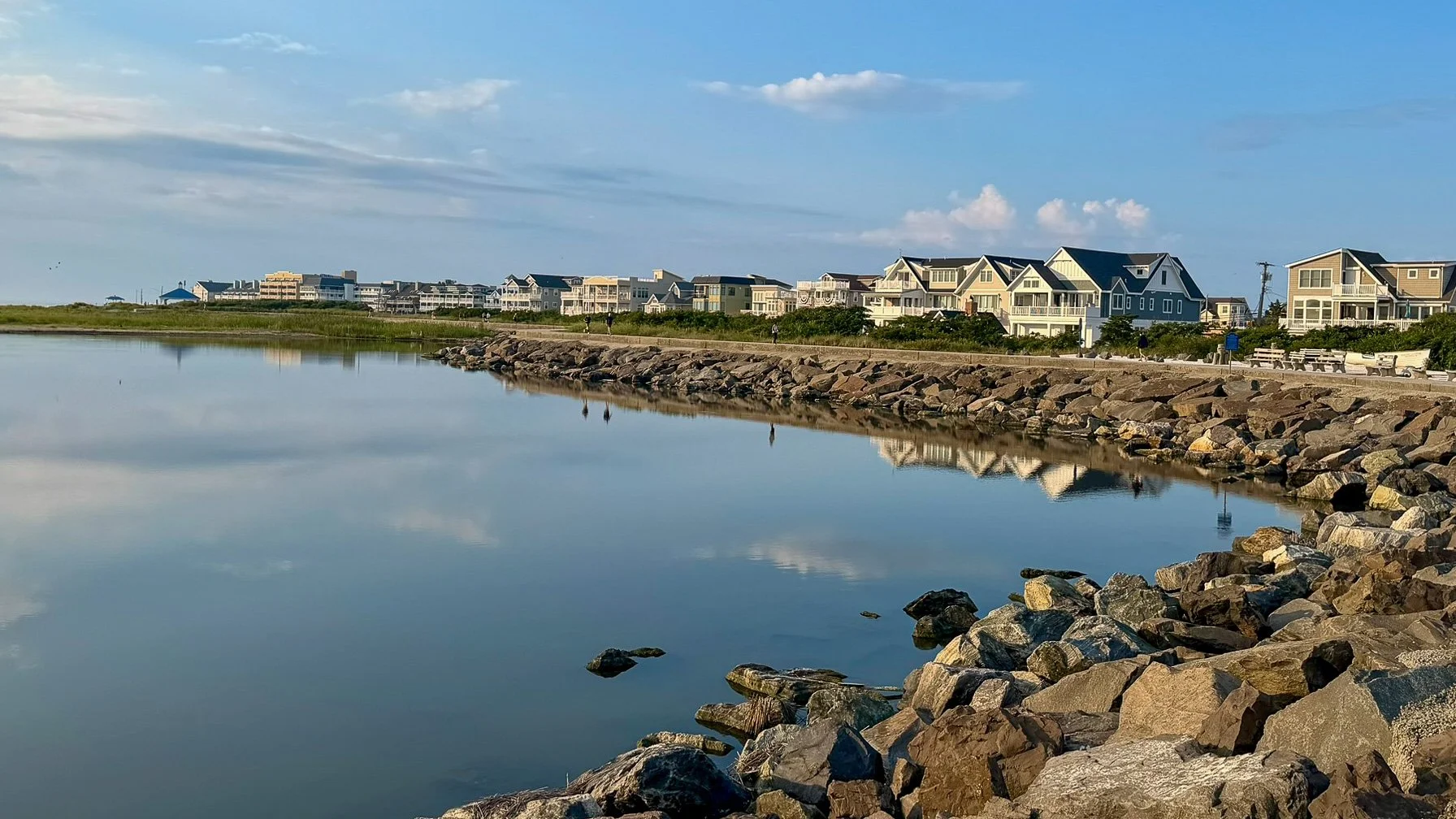 Calm lagoon in North Wildwood, New Jersey, reflecting waterfront homes and a blue morning sky beside a rocky shoreline.