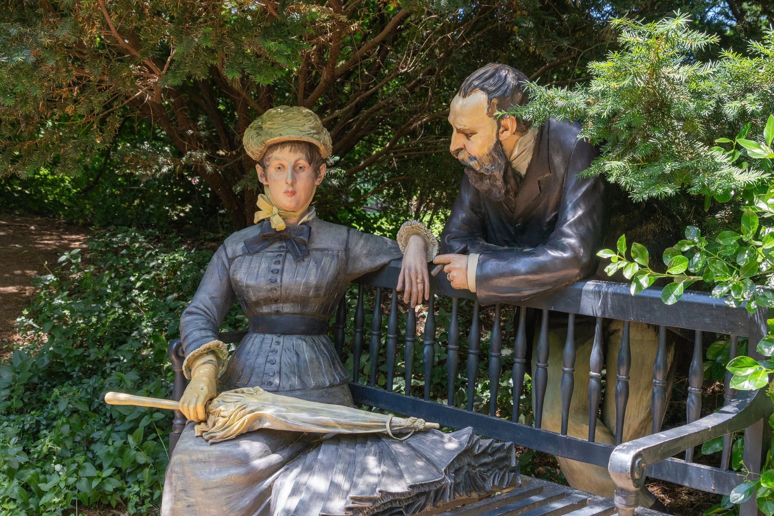 Victorian-era man and woman sculpture seated on a garden bench surrounded by trees at Grounds For Sculpture in Hamilton, New Jersey.