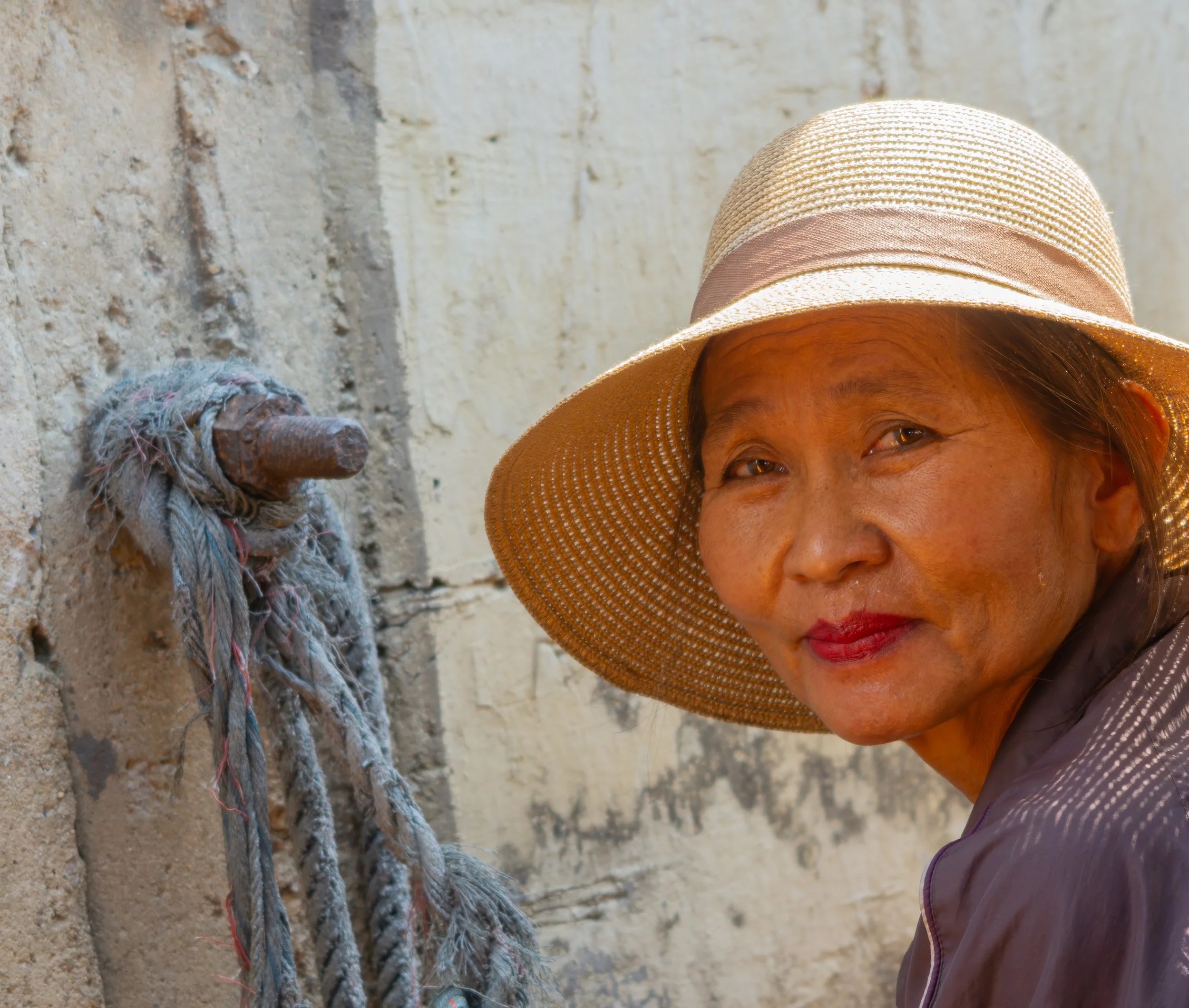 Thai woman wearing a woven sun hat looking back toward the camera beside a weathered wall.