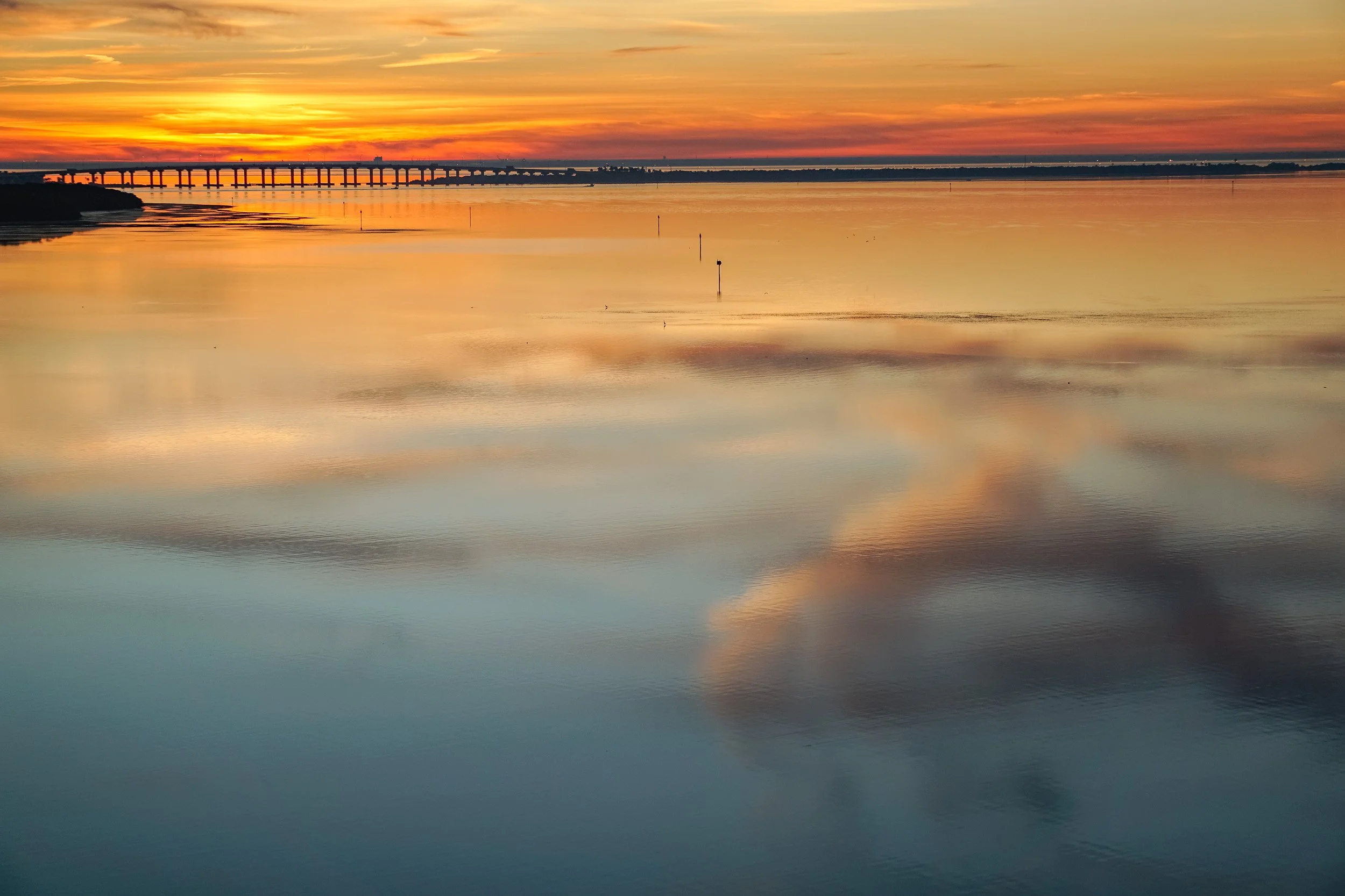 Golden sunset clouds reflected across the calm waters of Boca Ciega Bay in St. Petersburg, Florida, with a bridge silhouetted along the horizon.