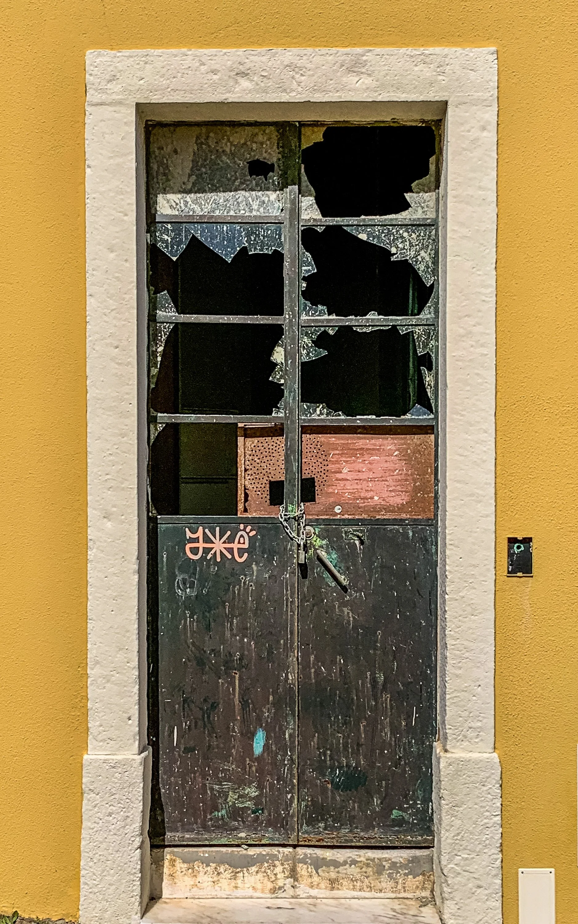 Weathered black metal doorway with broken windows set within a mustard-yellow wall in Portugal, revealing the textures of age, abandonment, and time.