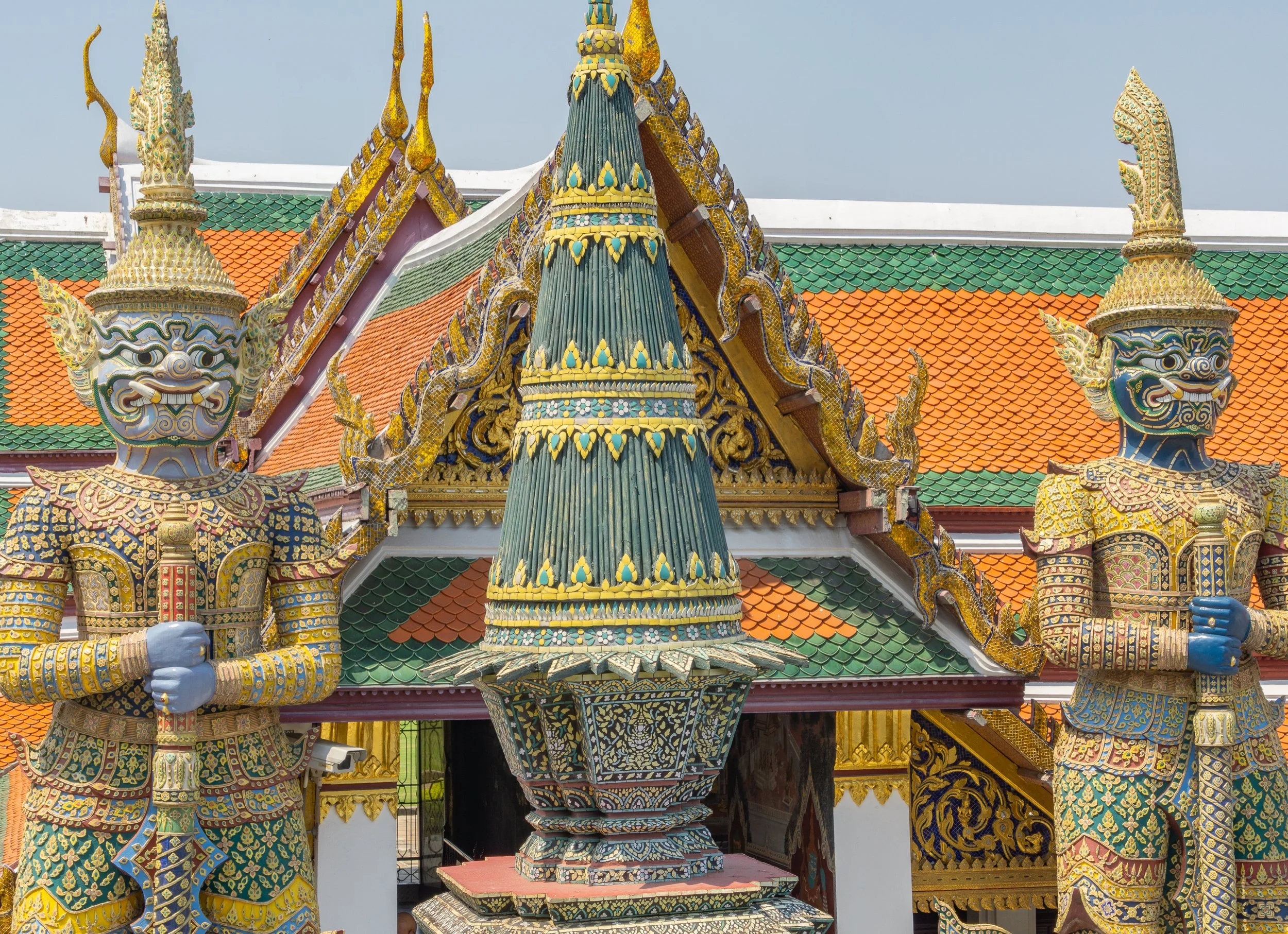 Colorful guardian statues stand at the entrance to the Grand Palace in Bangkok, Thailand, framed by ornate temple roofs and traditional Thai architecture.