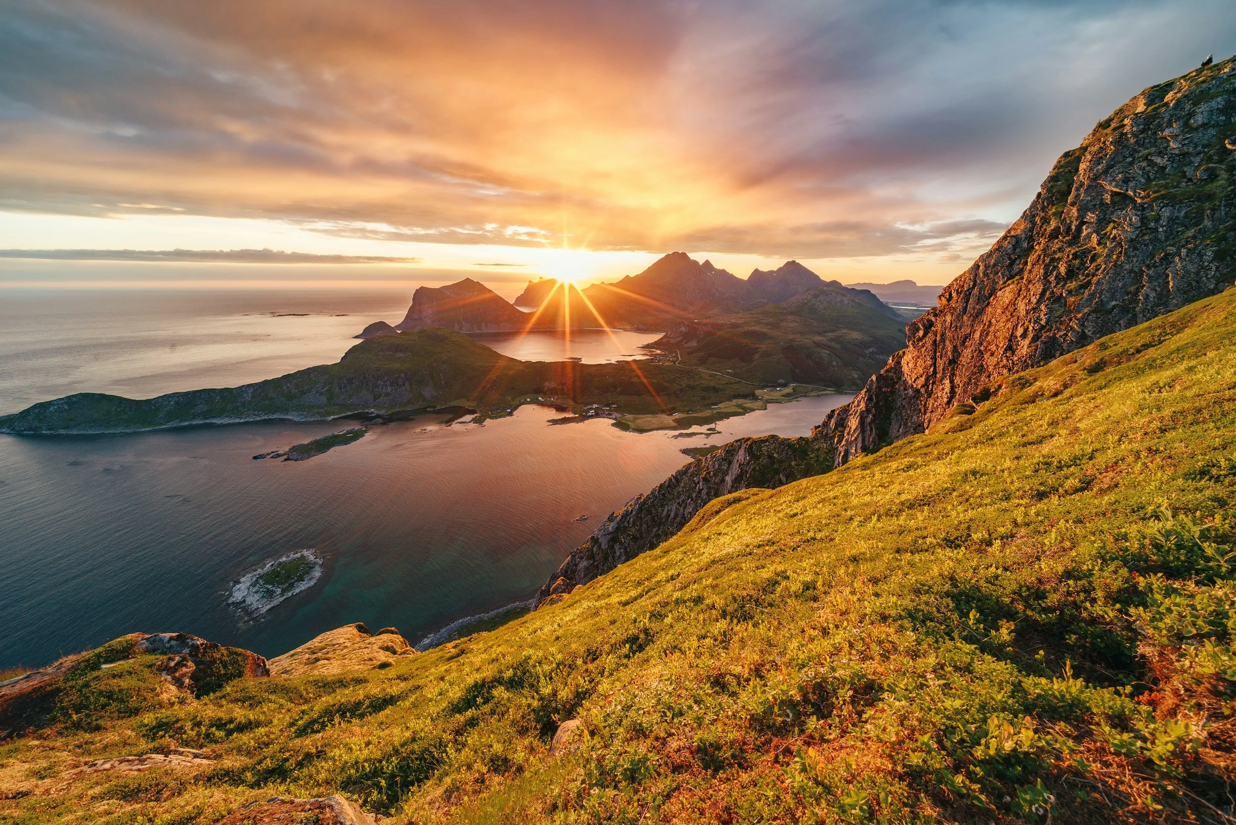 Sunrise over mountainous coastal landscape with water, rocky cliffs, and green vegetation.
