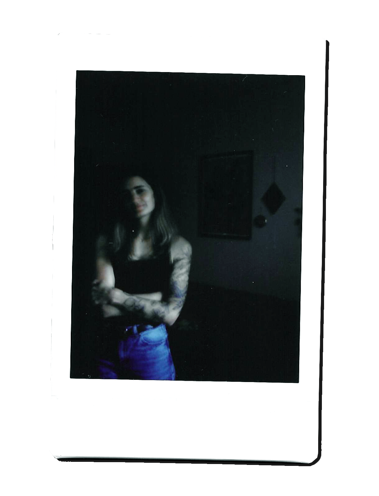 A woman with long hair and tattoos on her arms standing with arms crossed in a dimly lit room, wearing a black top and blue jeans.
