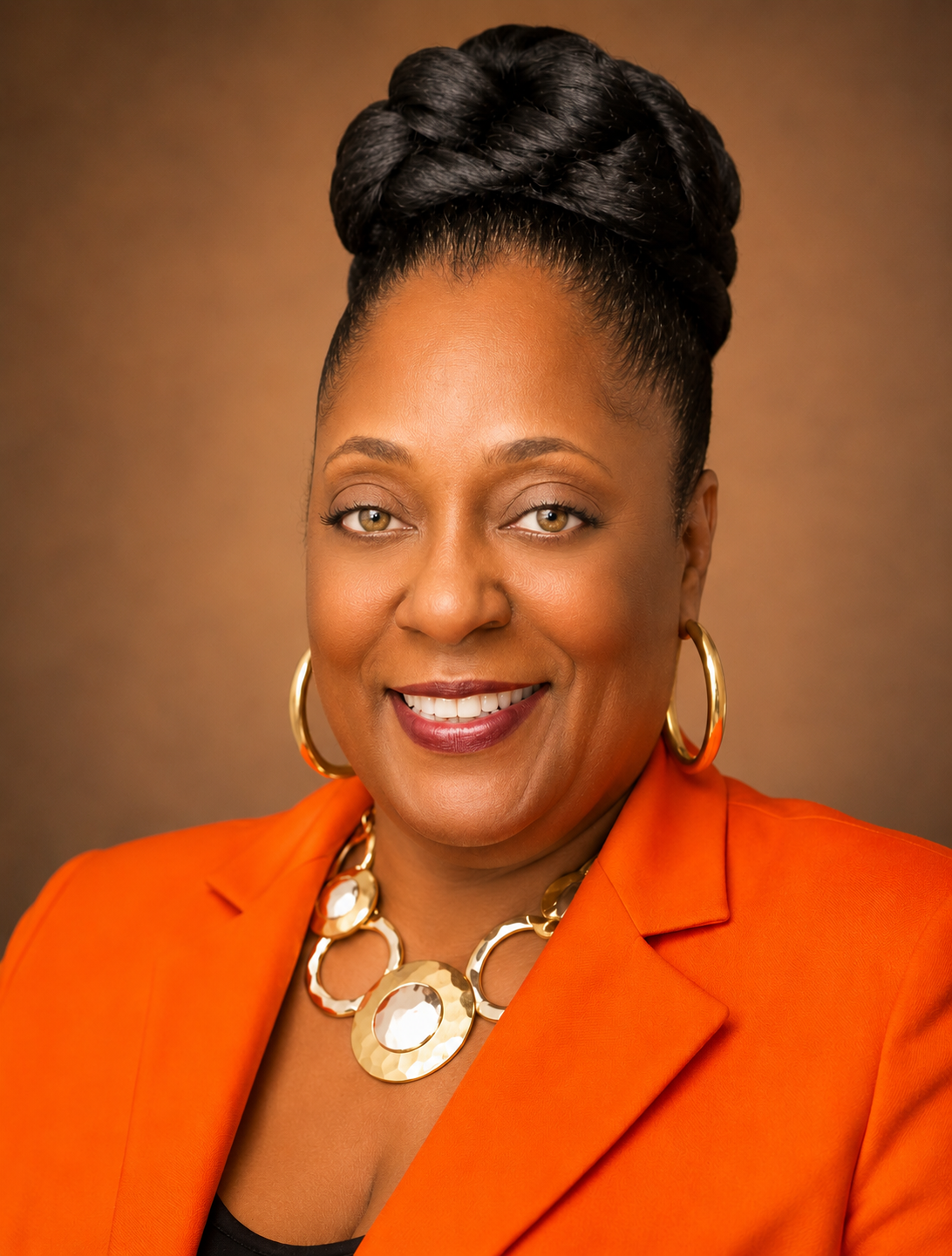 A smiling African American woman with braided hair styled in an updo, wearing a bright orange blazer, gold hoop earrings, and a large gold statement necklace, against a warm brown background.
