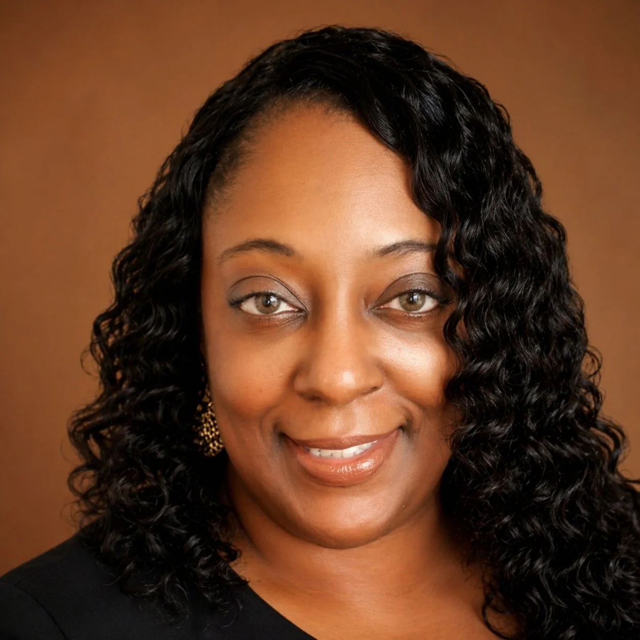 Close-up portrait of an African American woman with long, curly black hair, wearing gold earrings and a black top, smiling against a brown background.