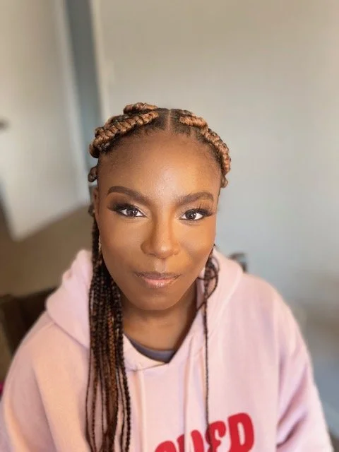 A woman with styled braids, wearing makeup and a pink hoodie, looking at the camera with a neutral expression in a well-lit room.