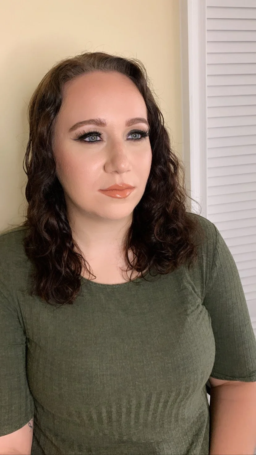 A woman with curly dark brown hair, blue eyes, and makeup including eyeliner, eyeshadow, and nude lipstick, looking to her left in a green top in front of a beige wall and louvered closet door.