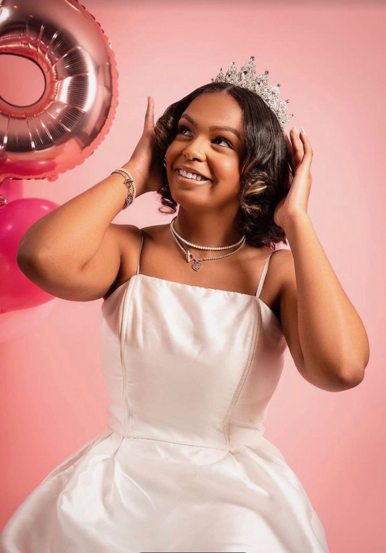 A woman wearing a white dress and pearl jewelry adjusts a silver tiara on her head in front of a pink background while smiling and holding a rose gold balloon.