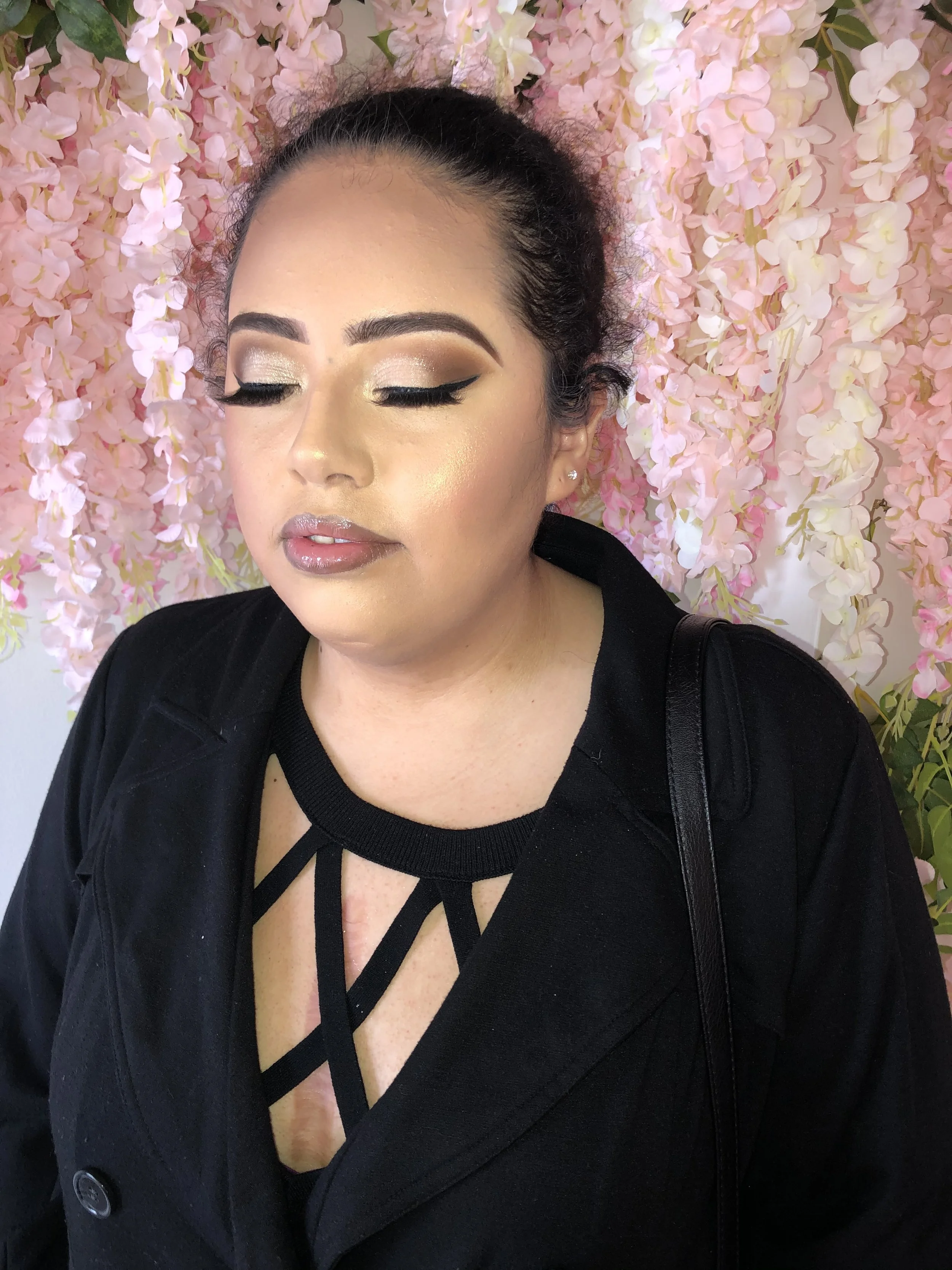 Close-up of a woman with makeup, closed eyes, and a black jacket, standing in front of a pink floral background.
