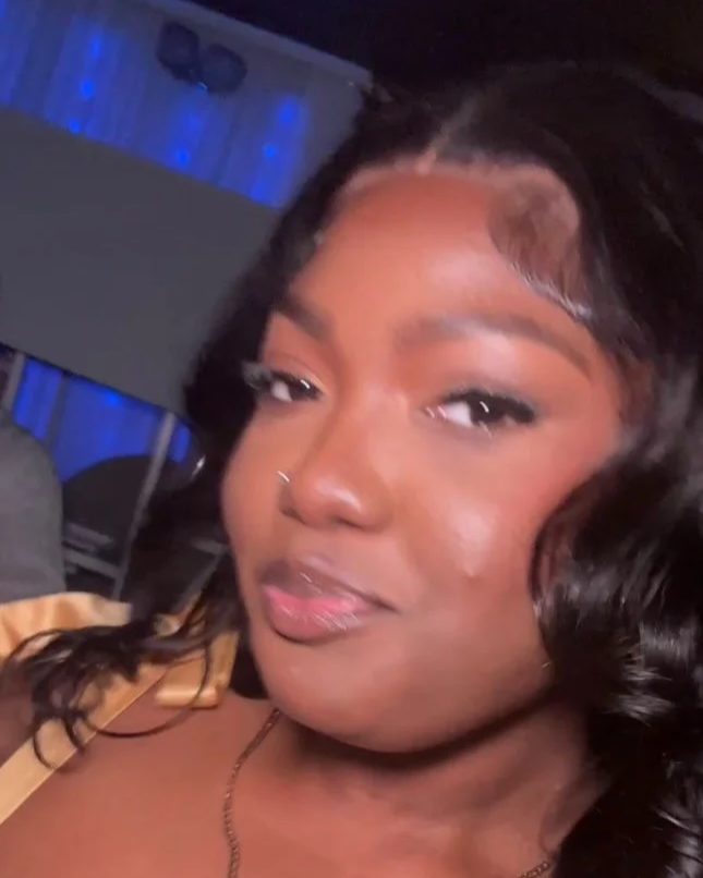 A woman with dark, highlighted hair and a nose piercing taking a selfie indoors at night with blue lighting in the background.