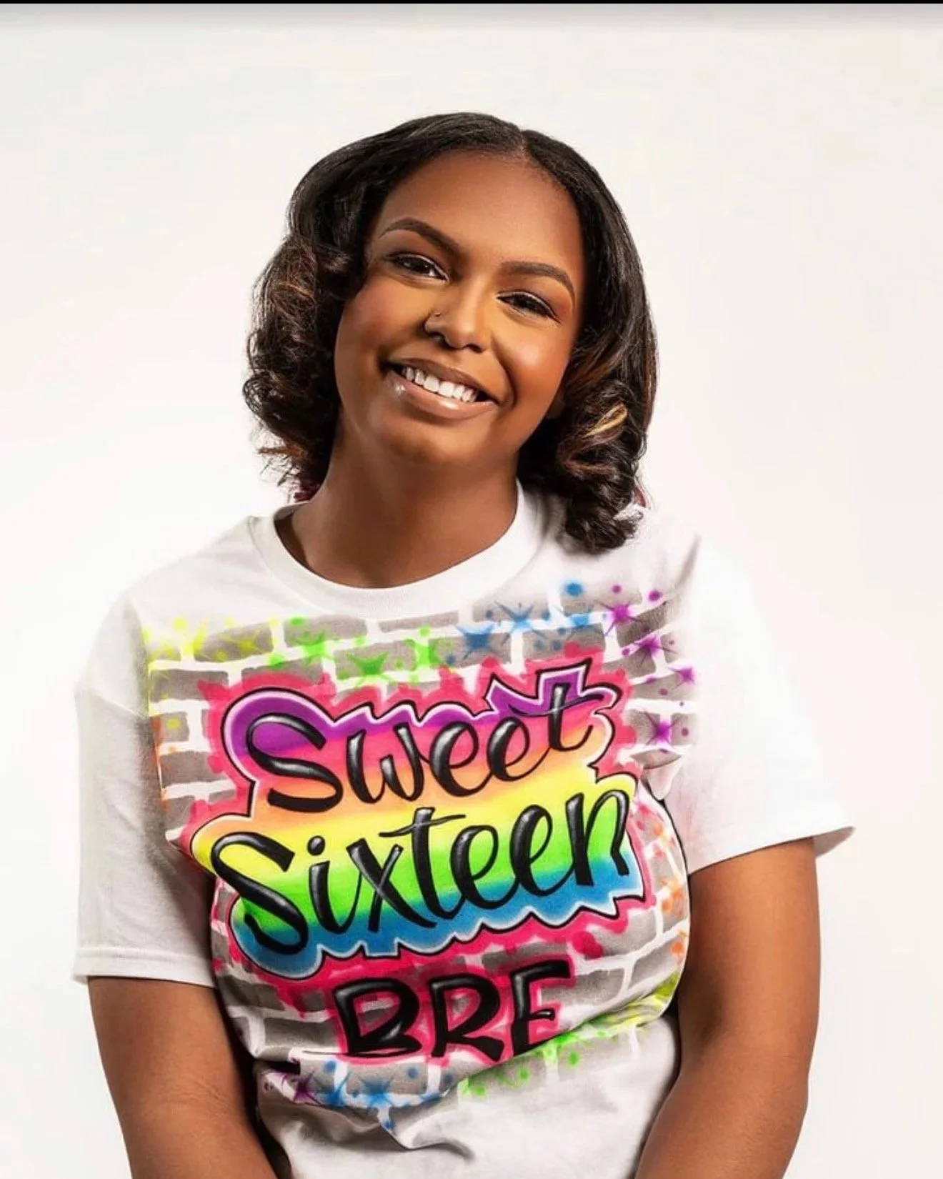 Young woman with dark hair styled in loose curls wearing a white T-shirt with colorful graffiti text that reads 'Sweet Sixteen B' against a plain white background
