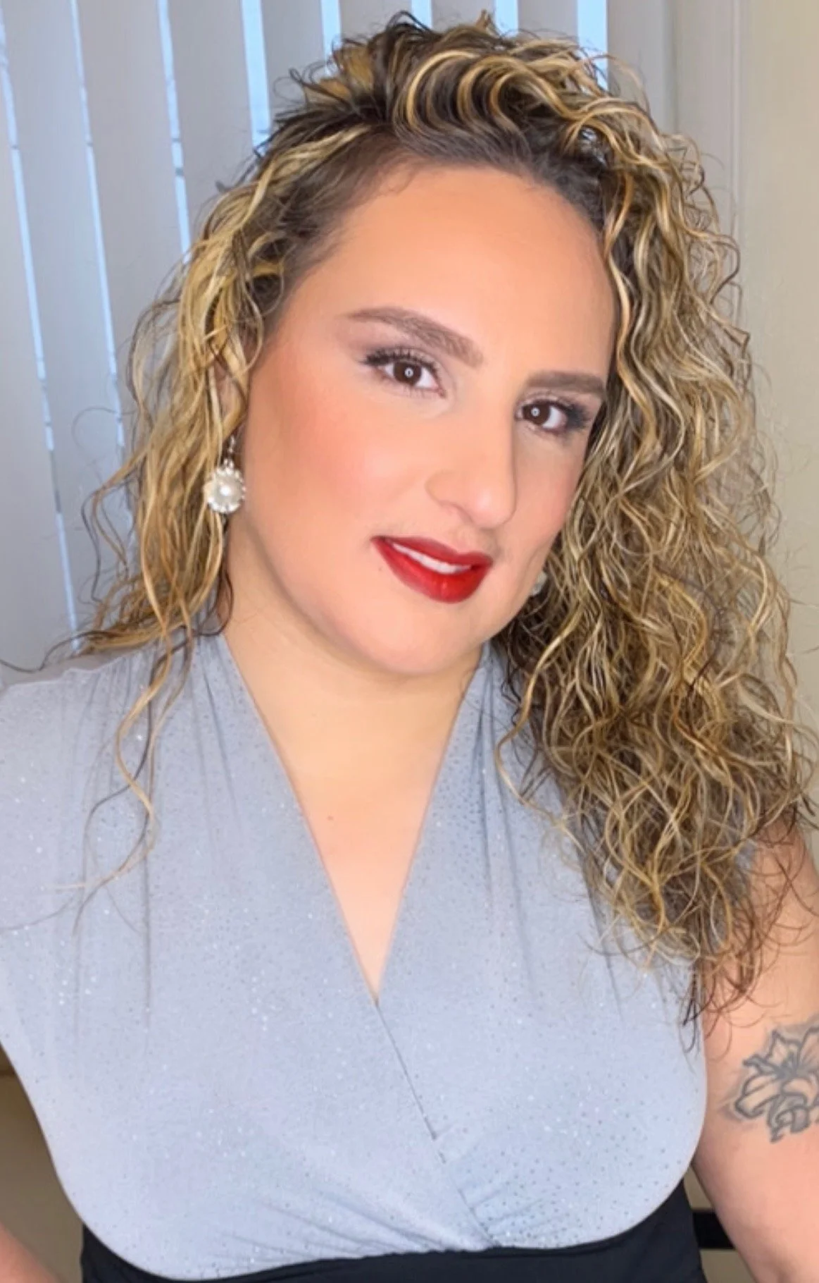 A woman with curly blonde hair wearing a light-colored top with earrings, standing in front of a background with vertical blinds.