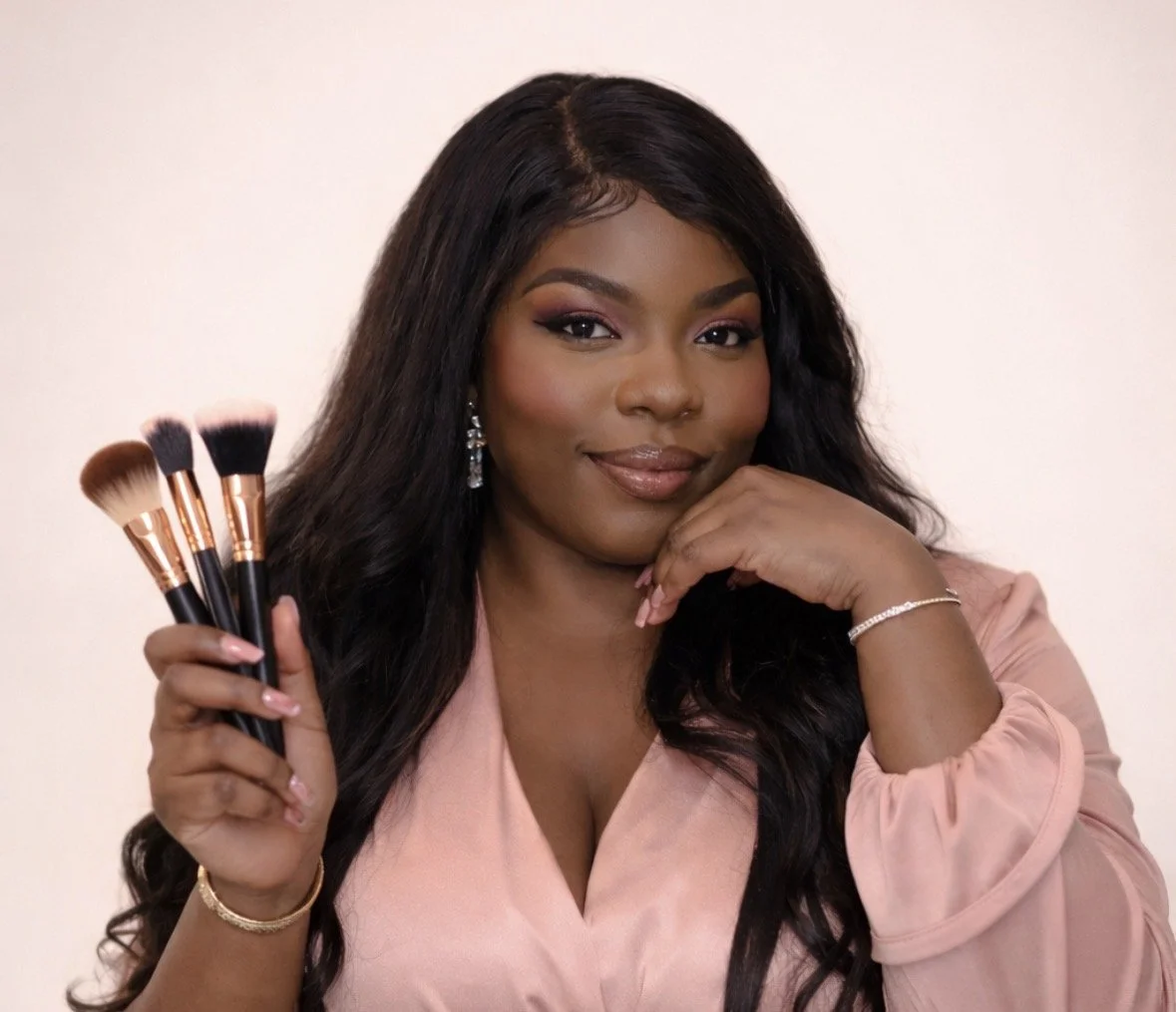A woman with long dark hair holding makeup brushes, wearing a pink top, and smiling at the camera.