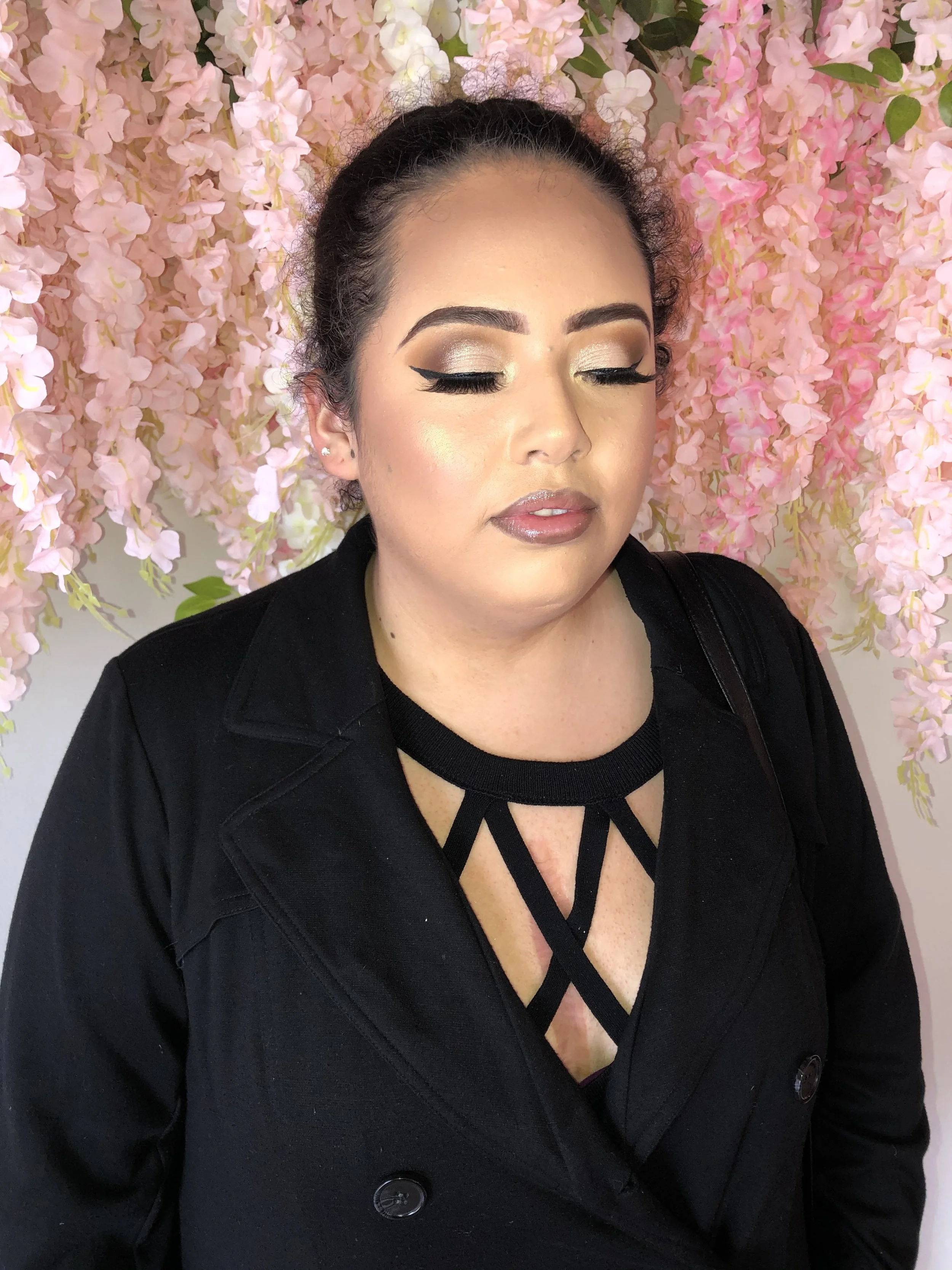 A woman with closed eyes and makeup, wearing a black outfit, standing in front of a pink floral backdrop.