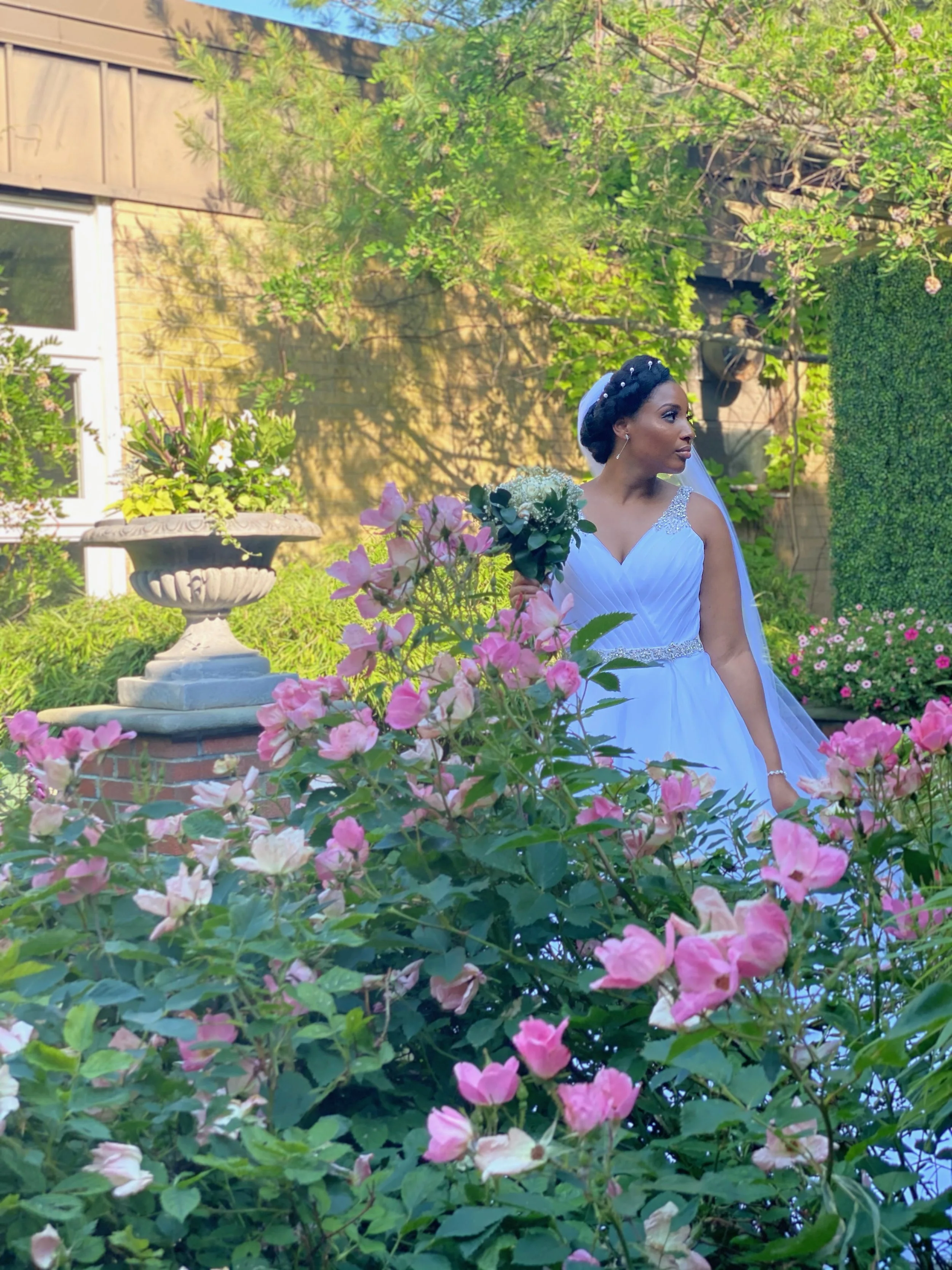 A bride in a white wedding dress holding a bouquet of white flowers in a garden with pink flowers and green foliage.