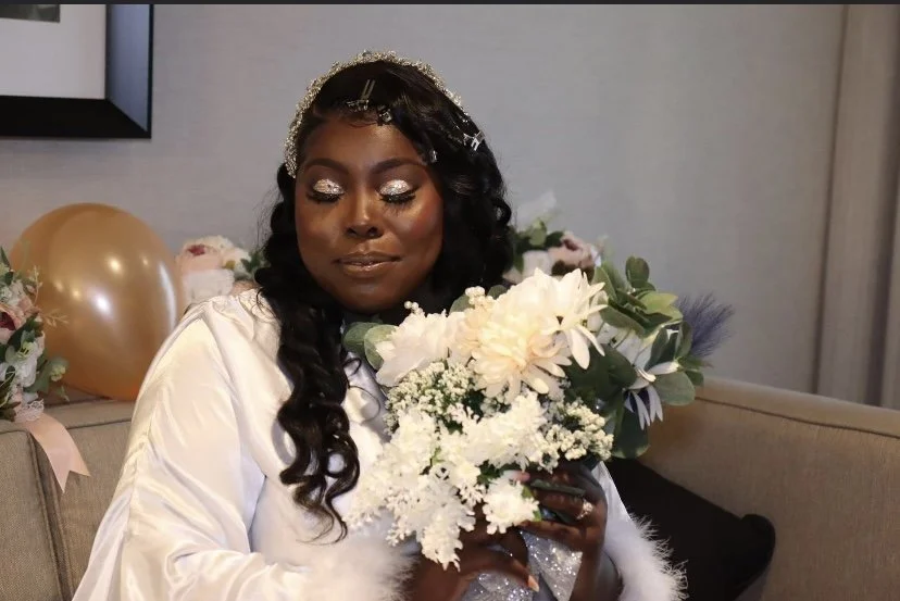 Woman with long dark curled hair holding a bouquet of white flowers in a room decorated with balloons and flowers, wearing a white satin robe with glitter makeup.