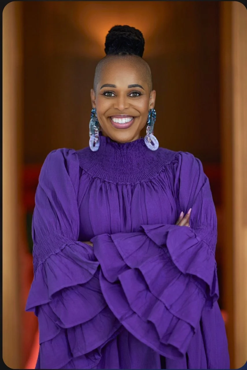 A smiling woman with a shaved head and large blue earrings, wearing a purple dress with ruffled sleeves, standing with arms crossed in front of a wooden background.