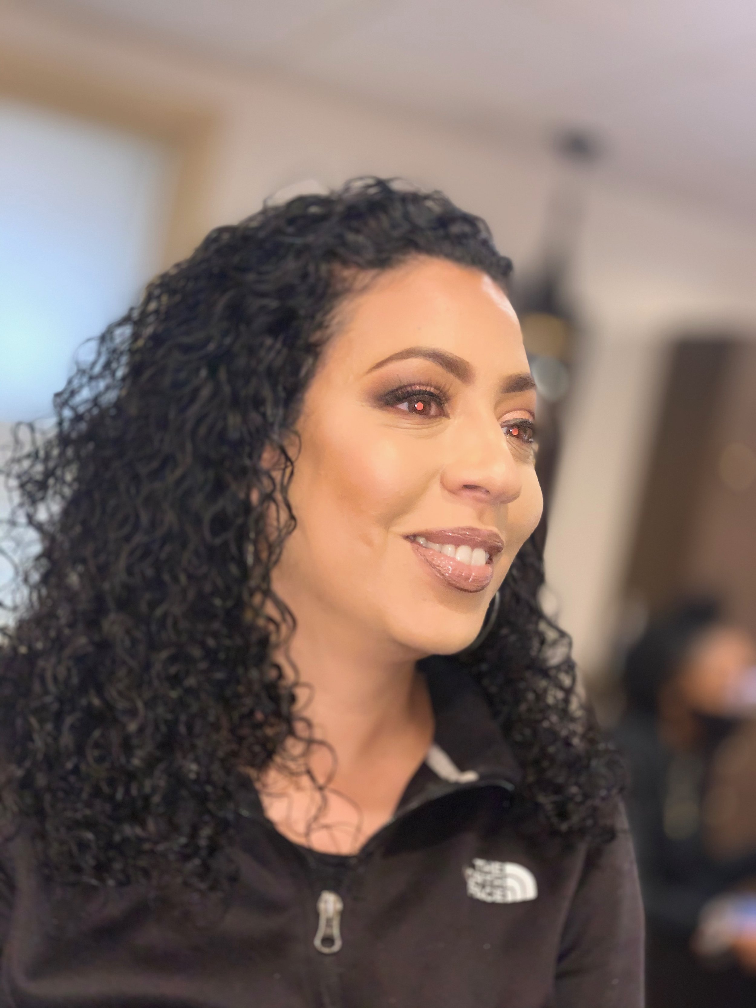 Close-up of a woman with dark curly hair, smiling, wearing makeup, and a black jacket, indoors.