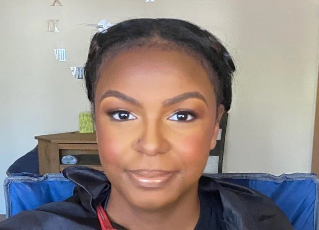 Close-up of a young woman with short braided hair smiling at the camera, wearing makeup, seated indoors with a clock and bookshelf in the background.