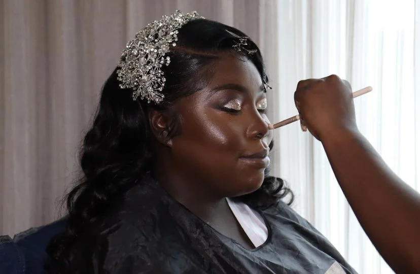 A woman with dark skin and black wavy hair being makeup finished, wearing a sparkling hair accessory and glitter eyeshadow, sitting with her eyes closed as someone applies makeup with a brush.