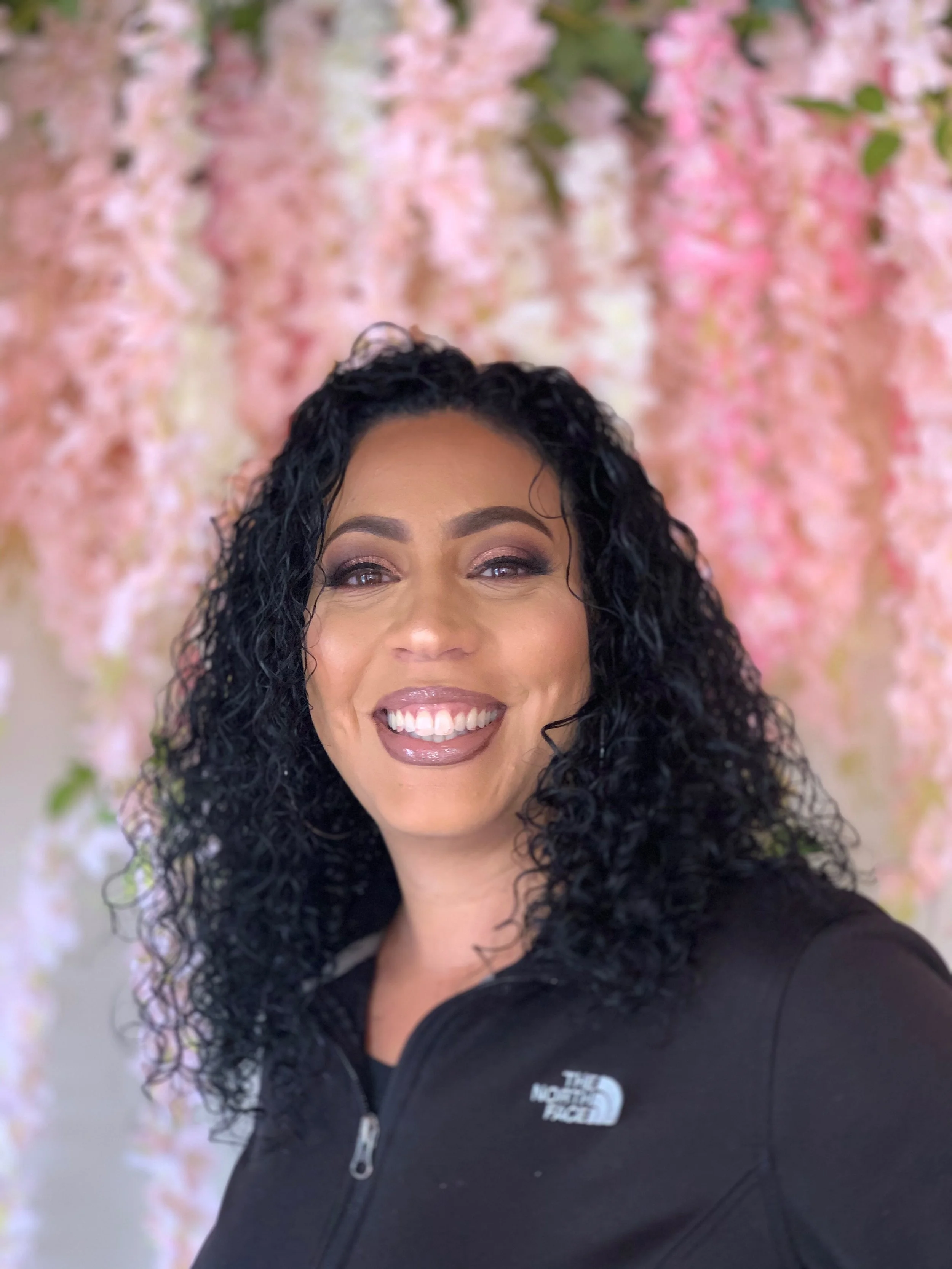 Close-up of a smiling woman with curly black hair, wearing makeup, in front of a pink floral background.