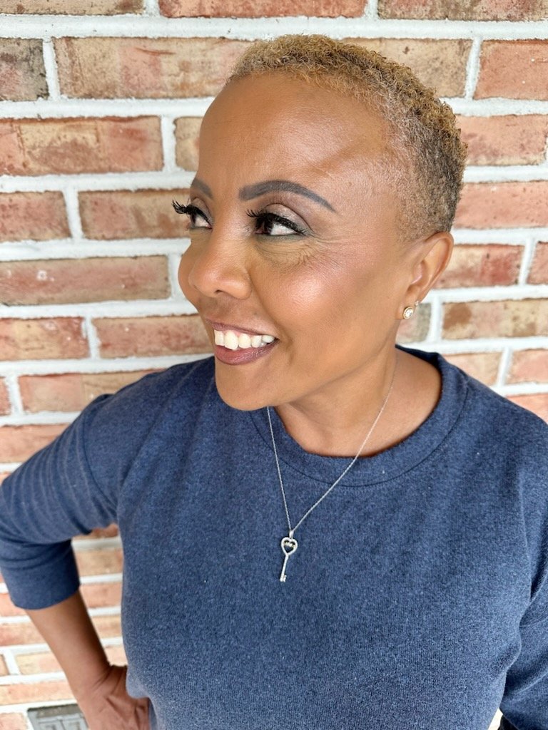 A woman with short, light brown hair and earrings, smiling and looking to her left. She is wearing a navy blue shirt, a necklace with a key pendant, and is standing in front of a brick wall.