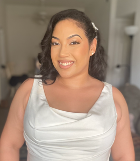 A woman with dark hair in loose curls, wearing a white sleeveless dress, smiling at the camera inside a home setting.