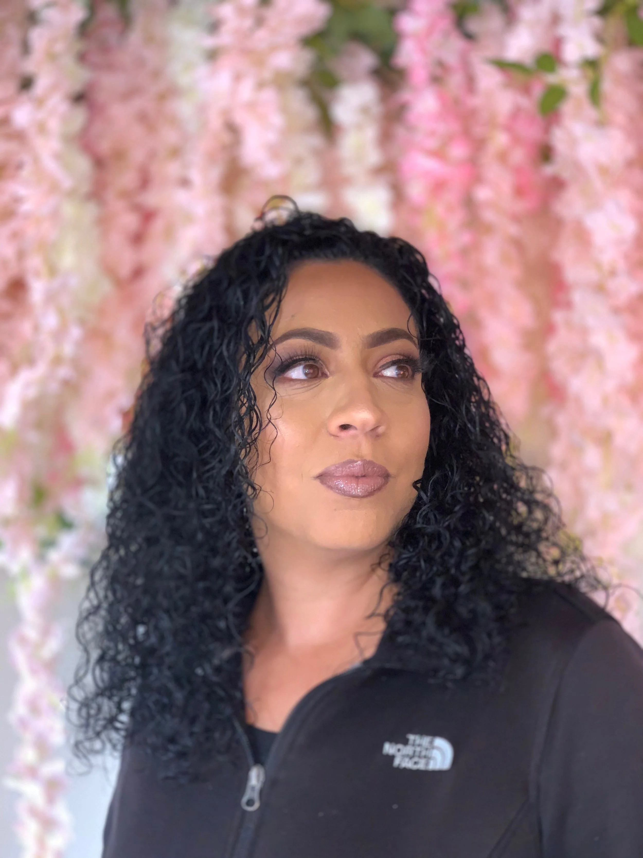 A woman with curly black hair and light brown skin, looking upwards, wearing a black North Face jacket, standing in front of a pink floral background.