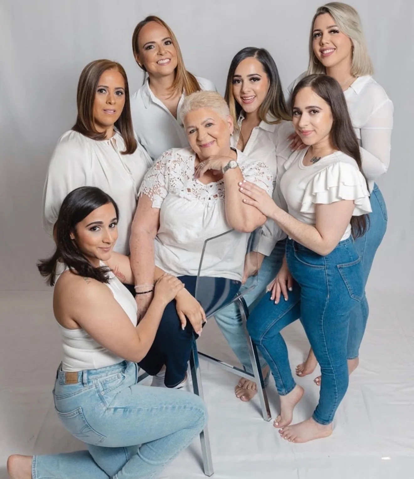 Group of women of various ages and skin tones posing together against a neutral backdrop, some standing and others sitting or kneeling, all wearing white tops and smiling at the camera.