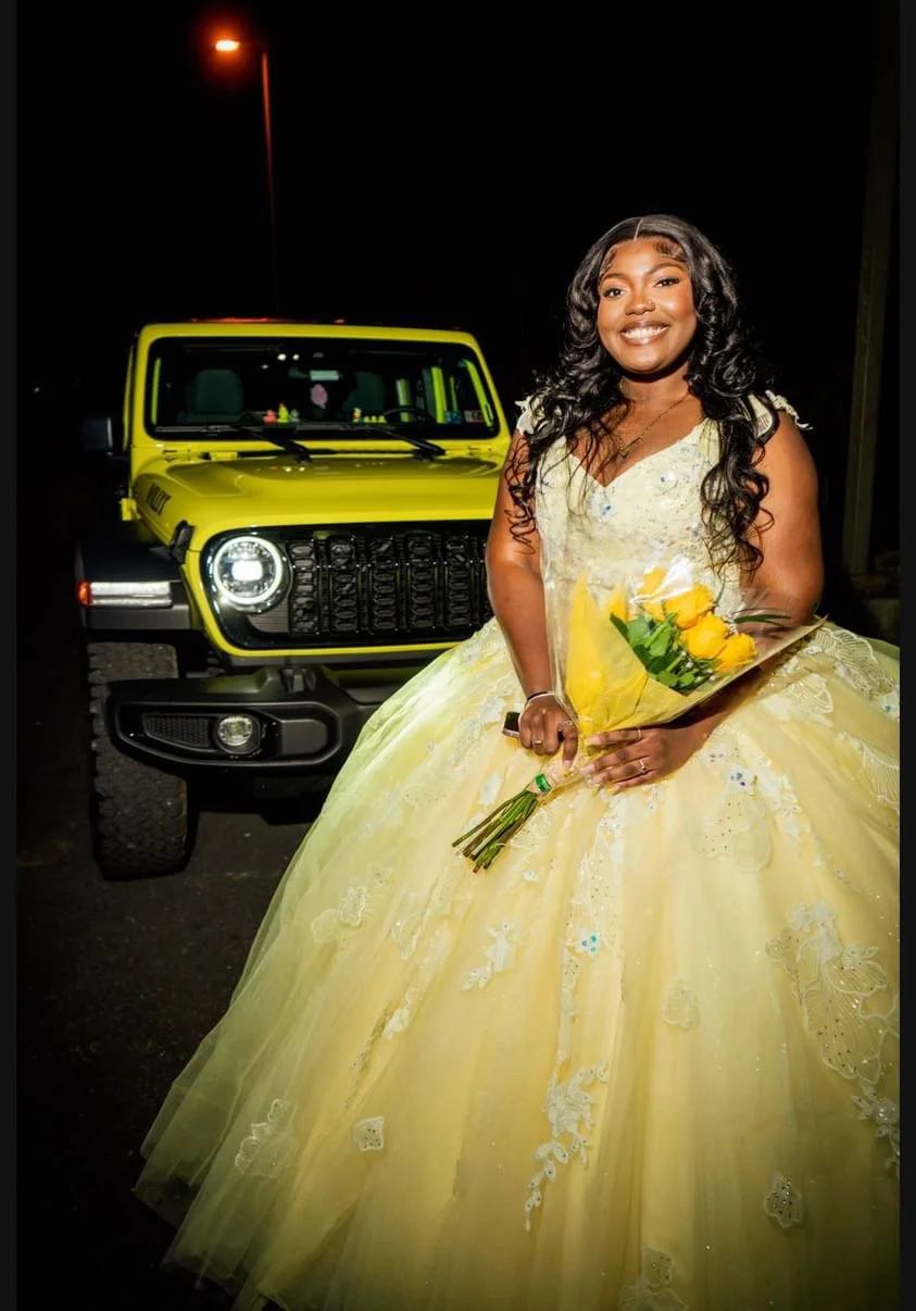 A young woman in a yellow gown holding a bouquet of yellow roses, standing in front of a bright yellow off-road vehicle at night, smiling.