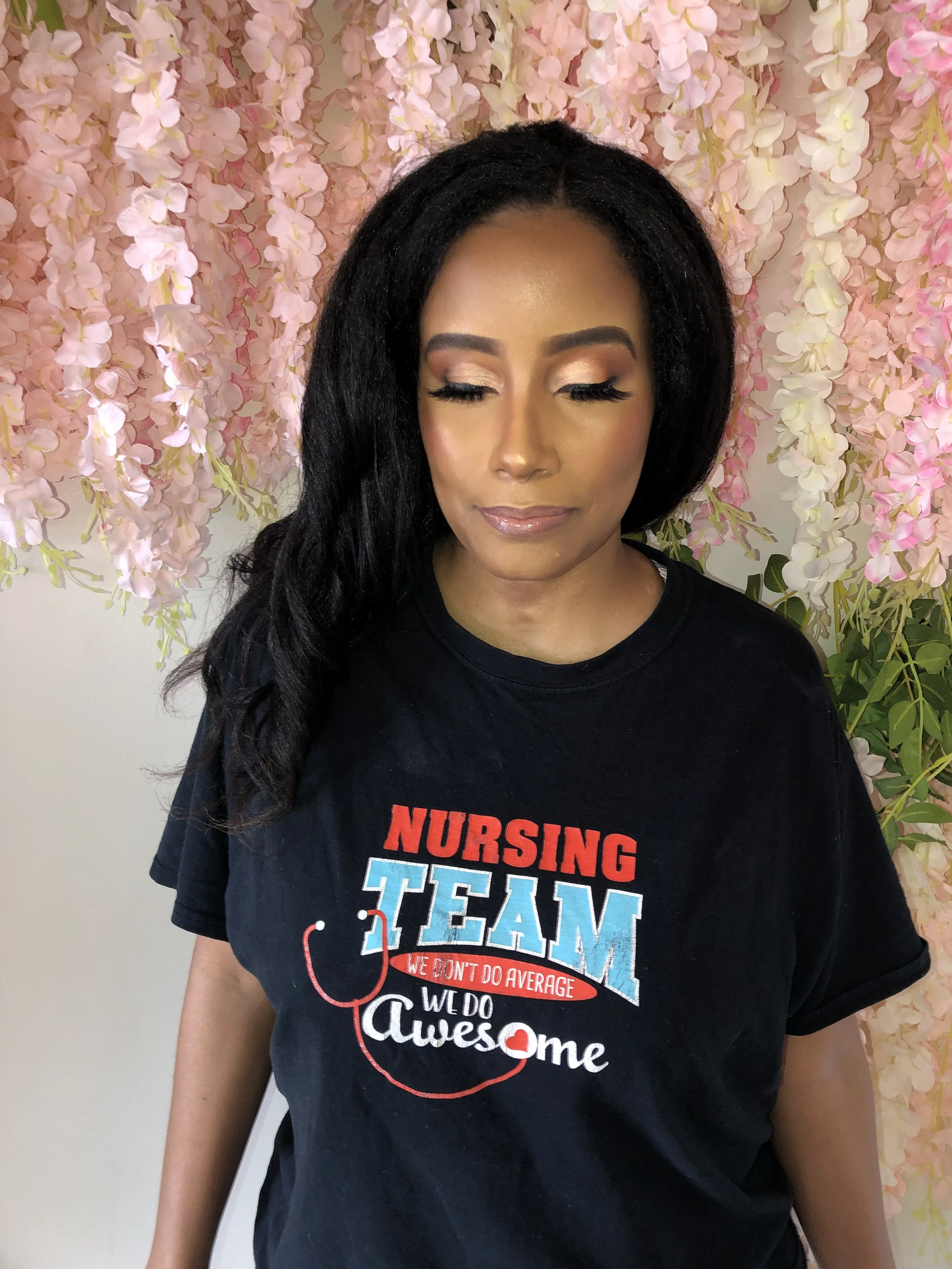 A woman with closed eyes and makeup, wearing a black T-shirt with red, white, and blue text and a stethoscope illustration. Pink floral backdrop.