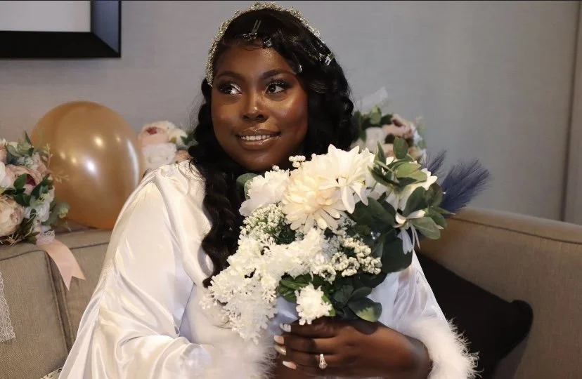 A woman with dark hair styled in loose curls holding a bouquet of white and green flowers, sitting on a beige sofa at a celebration with balloons and floral arrangements in the background.