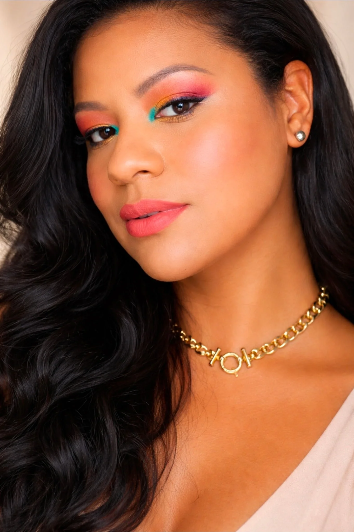Close-up of a woman with long dark hair, wearing colorful eye makeup, pink lipstick, and a gold necklace with the word "LOVE" on it, and a diamond stud earring.