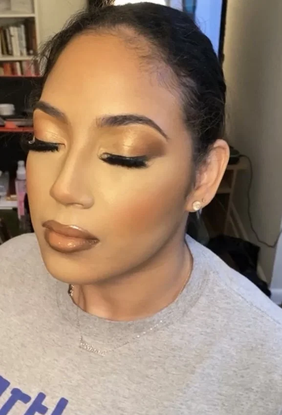 Close-up of a woman with makeup and her eyes closed, wearing a grey shirt and earrings, in a room with a bookshelf and a computer in the background.