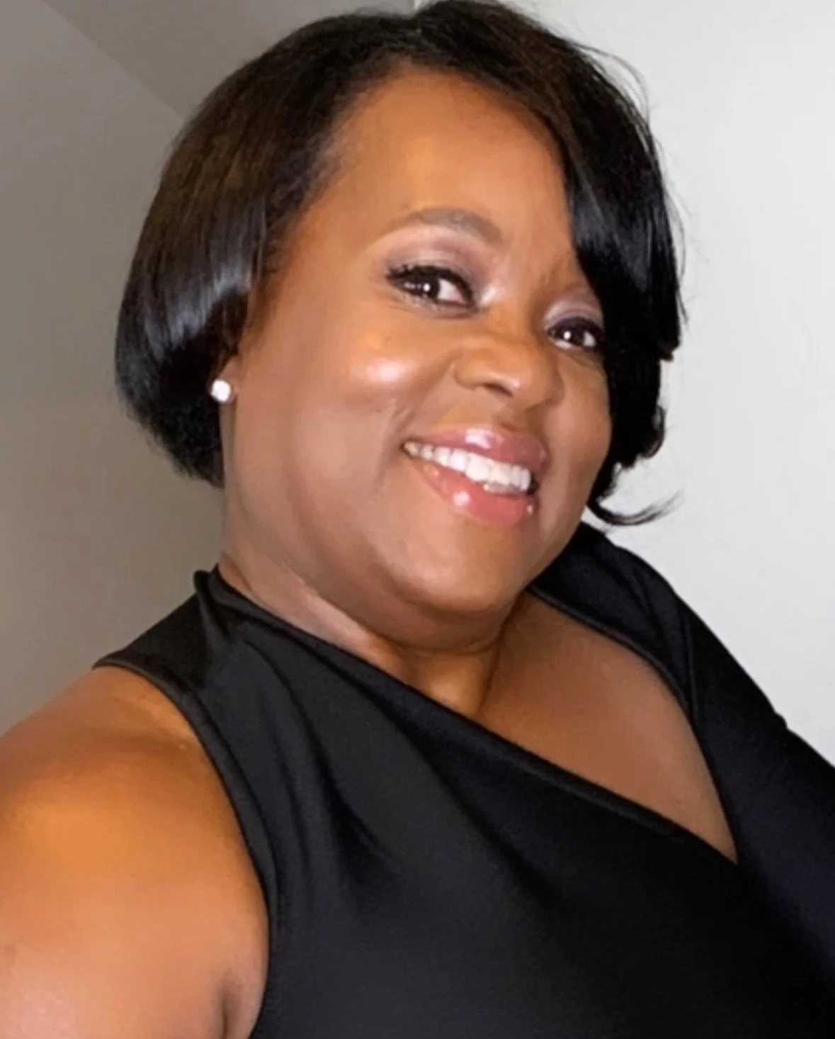 A woman with short black hair smiling, wearing pearl earrings and a sleeveless black top, standing indoors against a plain background.