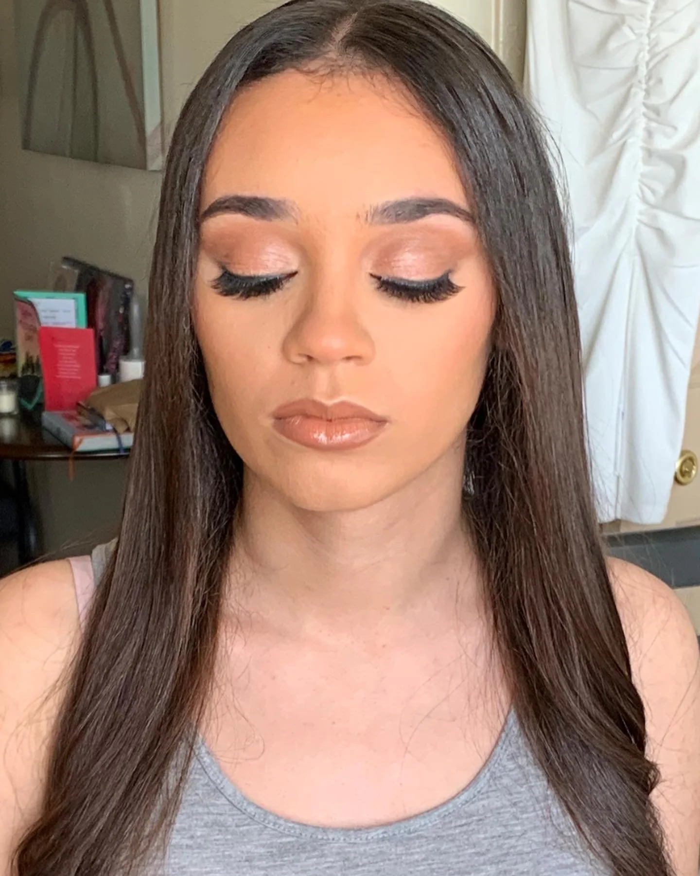 Close-up of a woman with long brown hair and closed eyes, wearing makeup including eyeshadow, mascara, and lipstick, in an indoor setting with various items in the background.