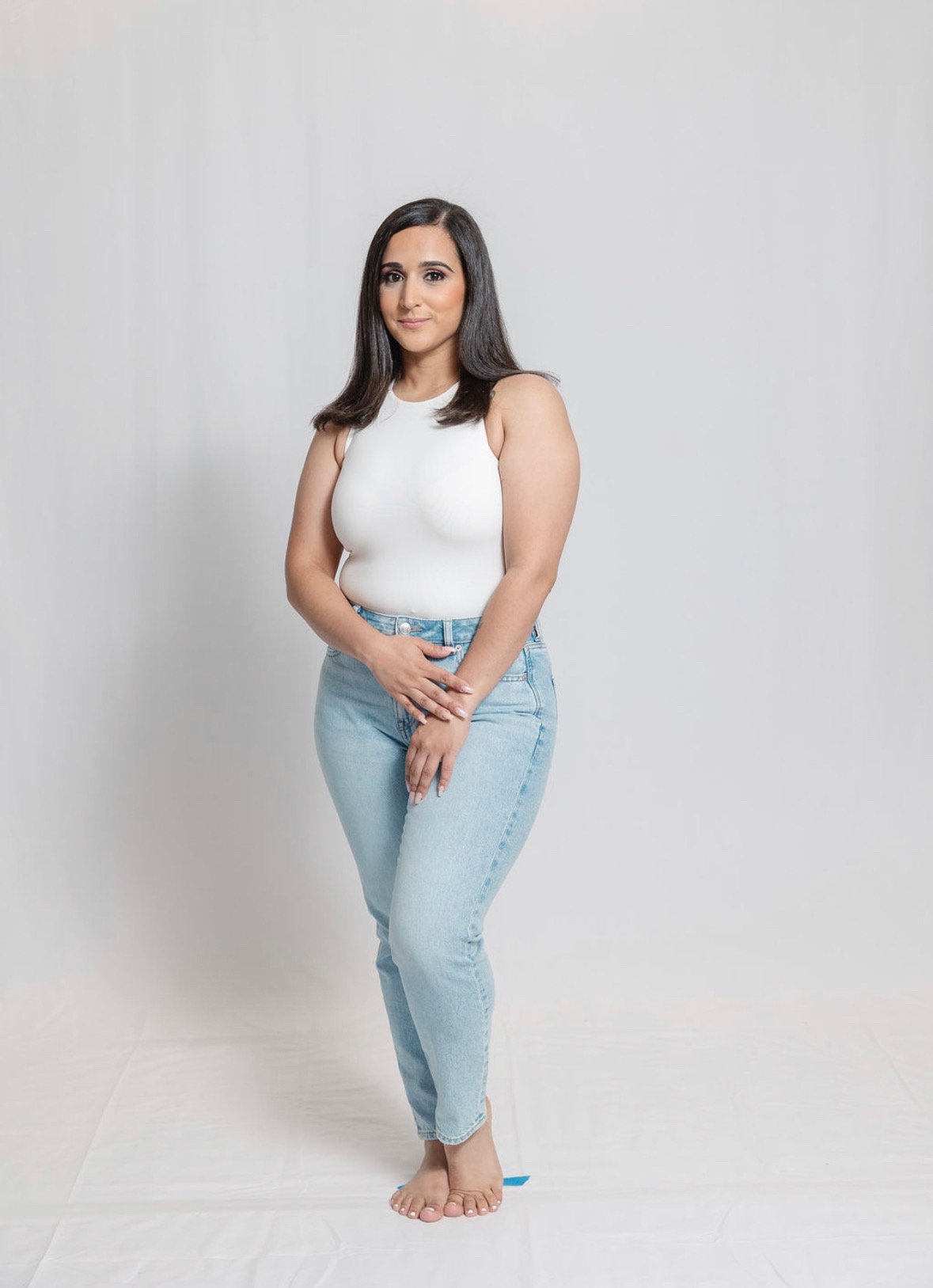 A woman with long dark hair, wearing a white sleeveless top and light blue jeans, standing against a plain white background, facing camera with a slight smile.
