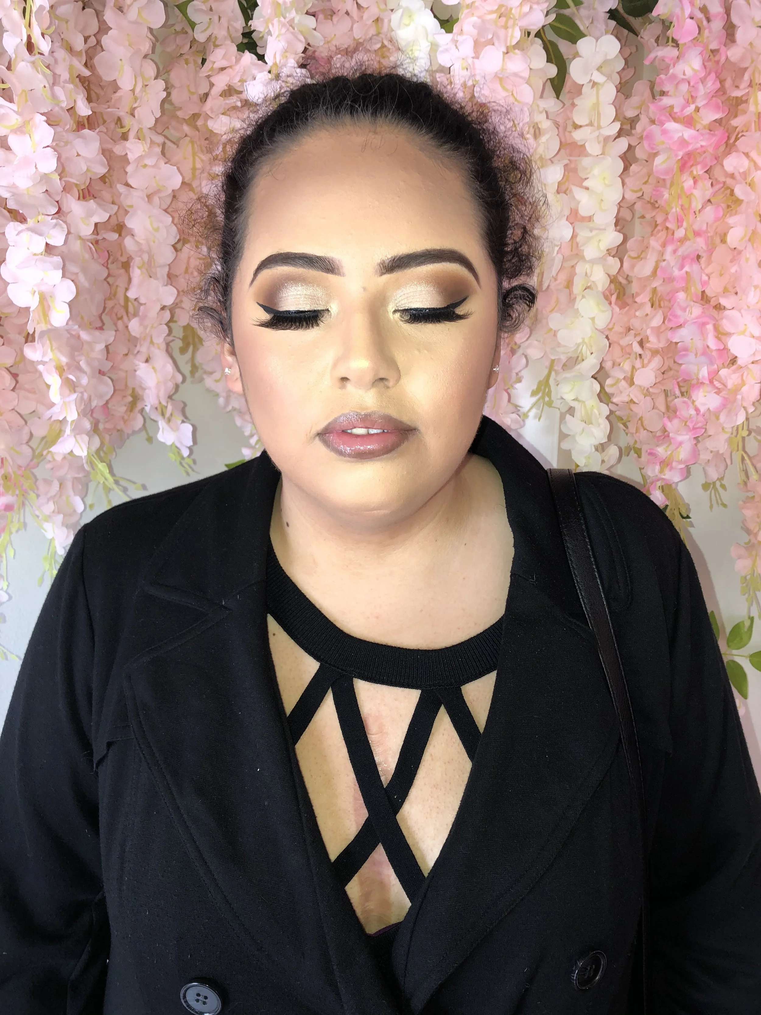 A woman with her eyes closed, wearing makeup and a black top with a crisscross neckline, standing in front of a pink floral background.