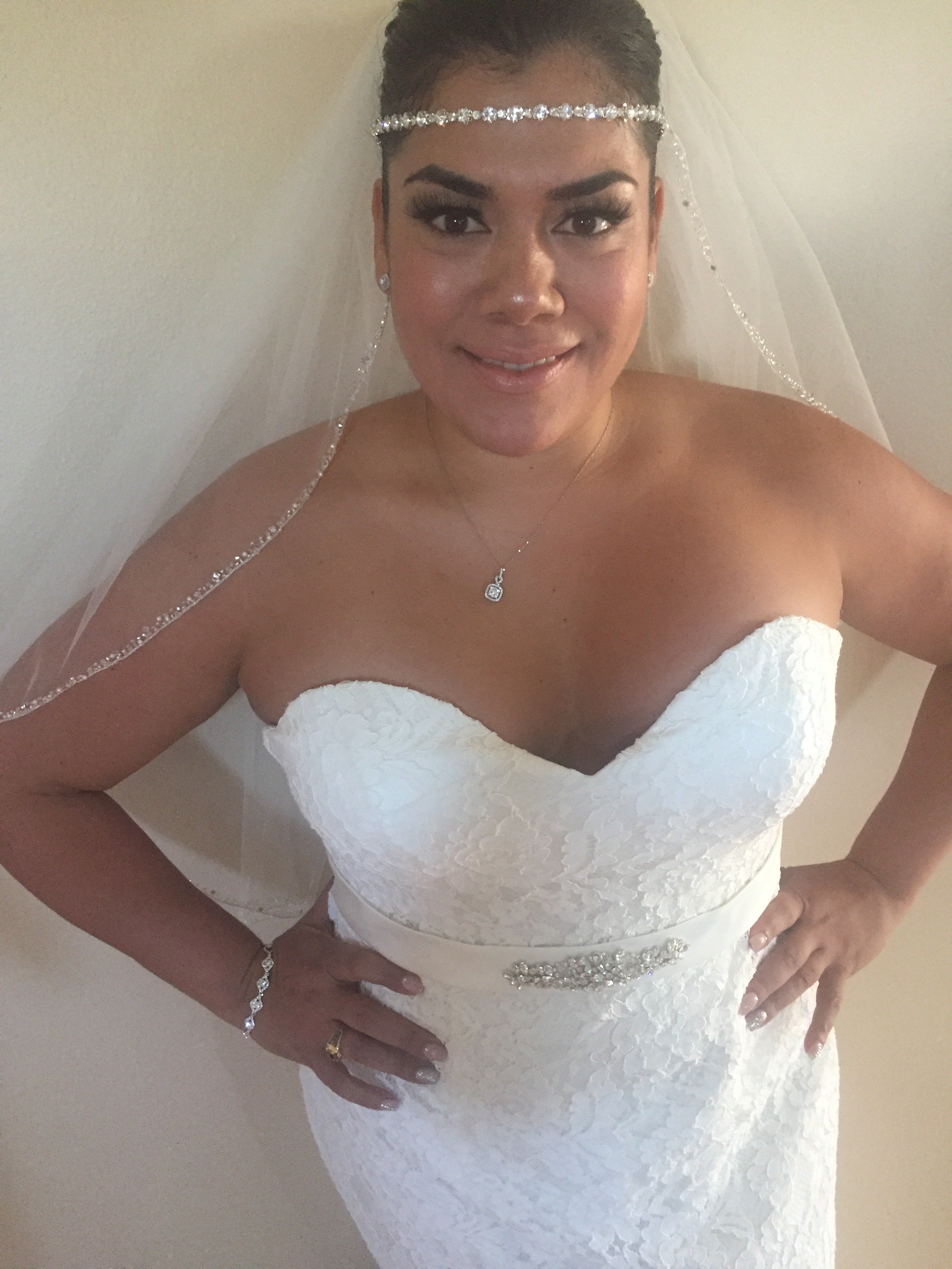 A woman dressed in a white lace wedding gown with rhinestone embellishments, wearing a veil, a diamond necklace, earrings, and a bracelet, smiling for the camera.