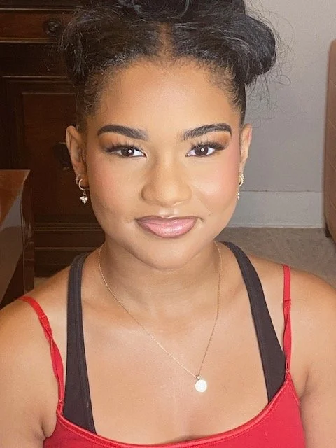 A young woman with dark curly hair styled in an updo, wearing makeup, small earrings, a necklace with a round pendant, a red and black tank top, smiling indoors.