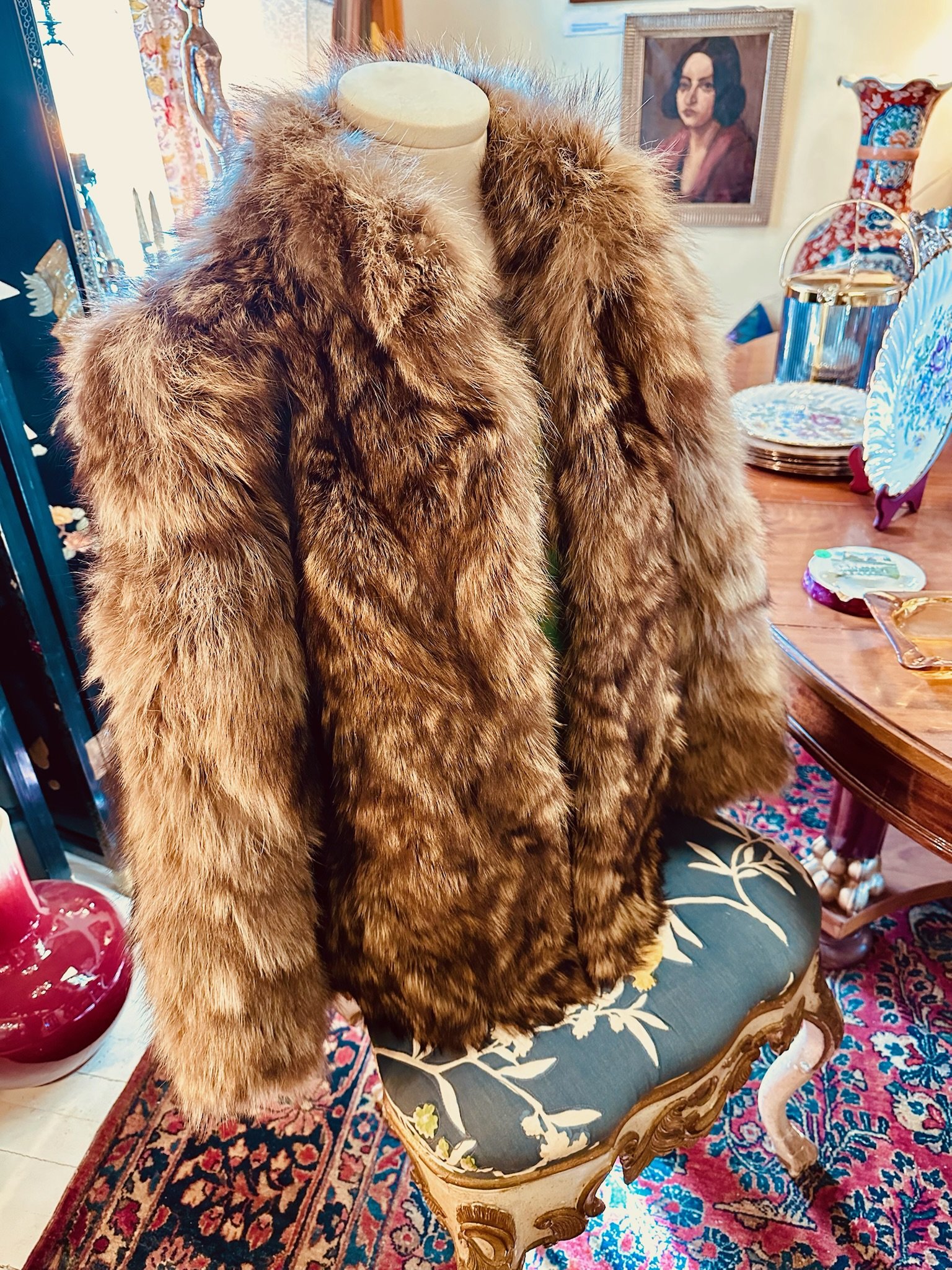 A customer consigned vintage raccoon fur coat for sale at Villa Vintiques. The background shows a wooden dining table with decorative plates and a teapot, and a framed portrait of a woman hanging on the wall.