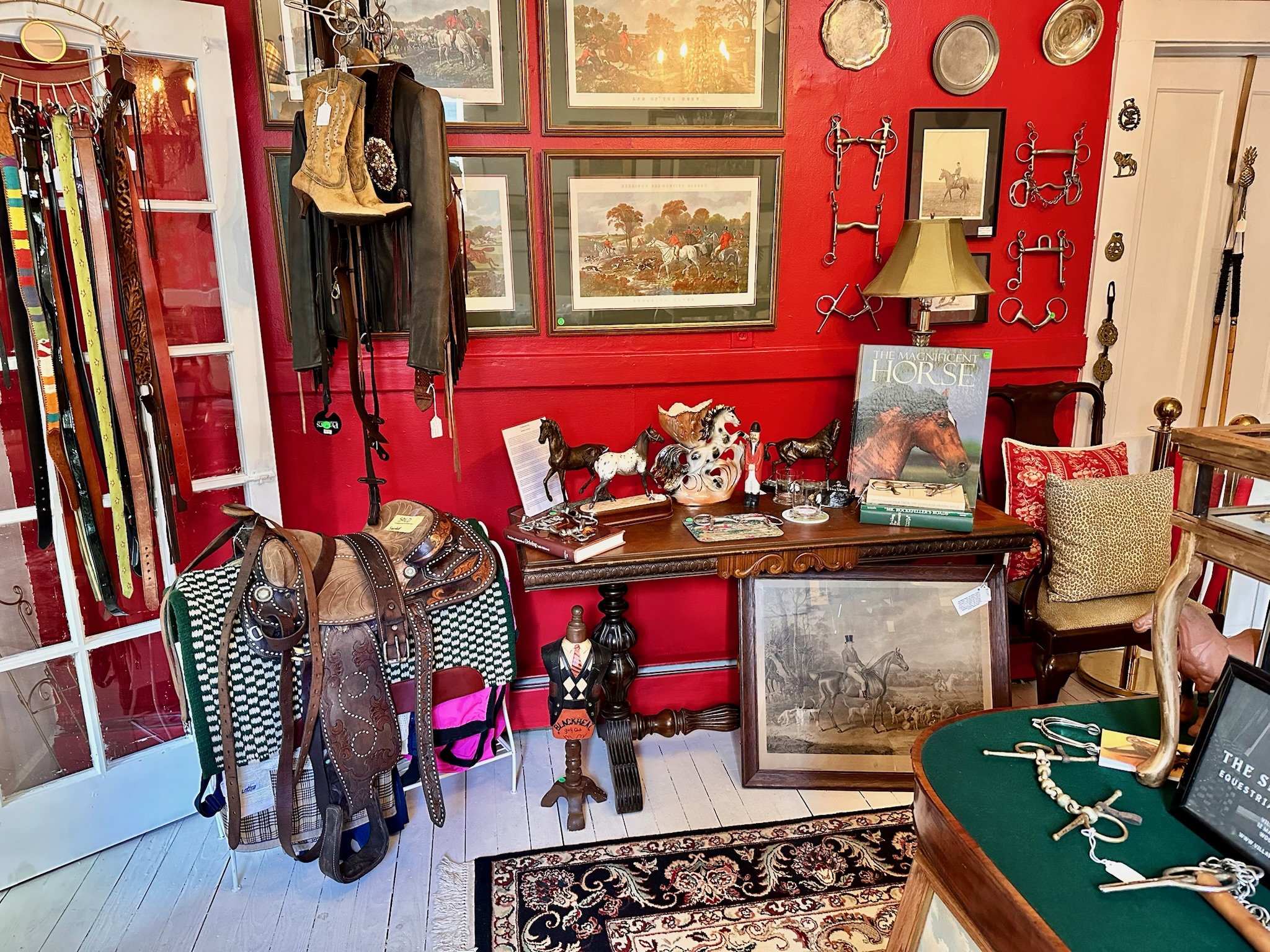 The Silver Spur Equestrian Consignment room at Villa Vintiques. Interior is decorated in Ralph Lauren Dressage red paint, with framed pictures, shelves of riding gear, horse models, and various antiques including silver trays and jewelry.