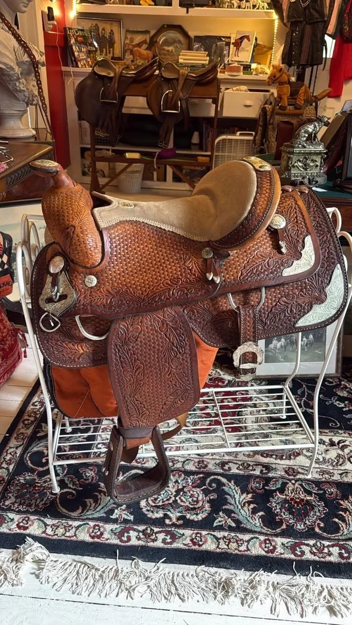 Leather western saddle with detailed tooling and silver accents placed on a white metal rack inside a store, with various cowboy and western-themed items in the background.