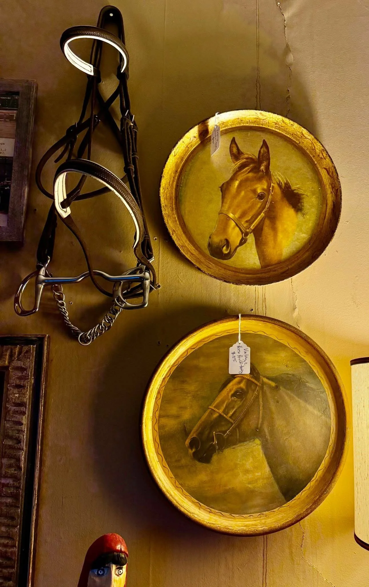 Decorative wall art of two oval wooden plaques featuring painted portraits of brown horses, with a horse riding bridle hanging on the wall to the left.