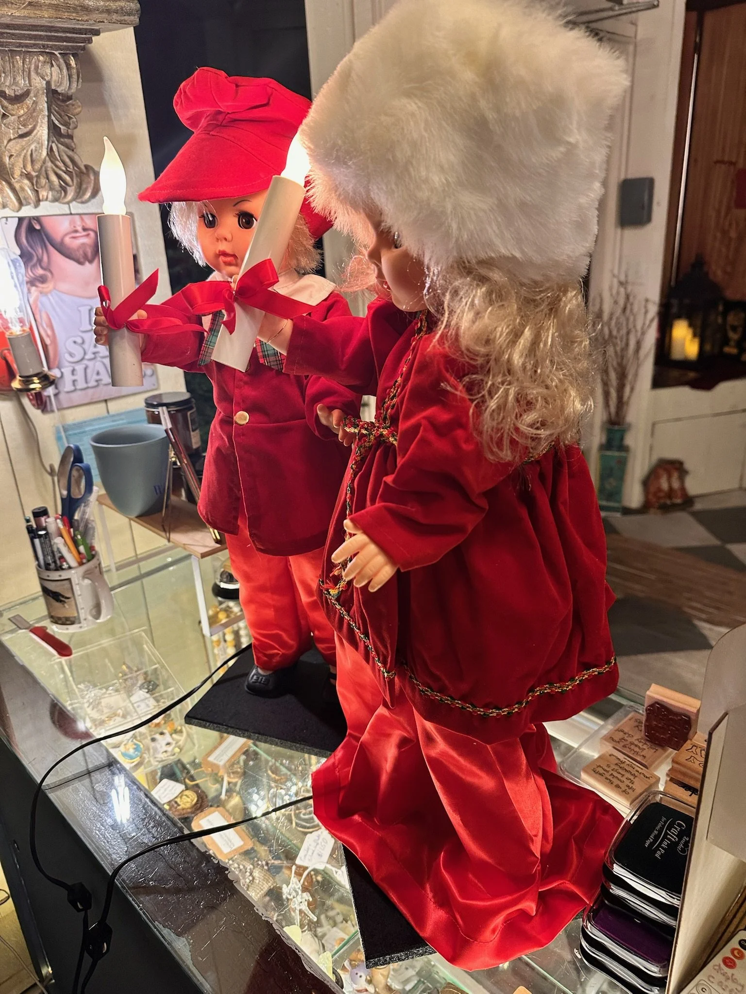 Vintage holiday decor at Villa Vintiques. Two dolls dressed in red with white fluffy hats and one with a red hat with a bow, standing behind a glass display case filled with jewelry and accessories.