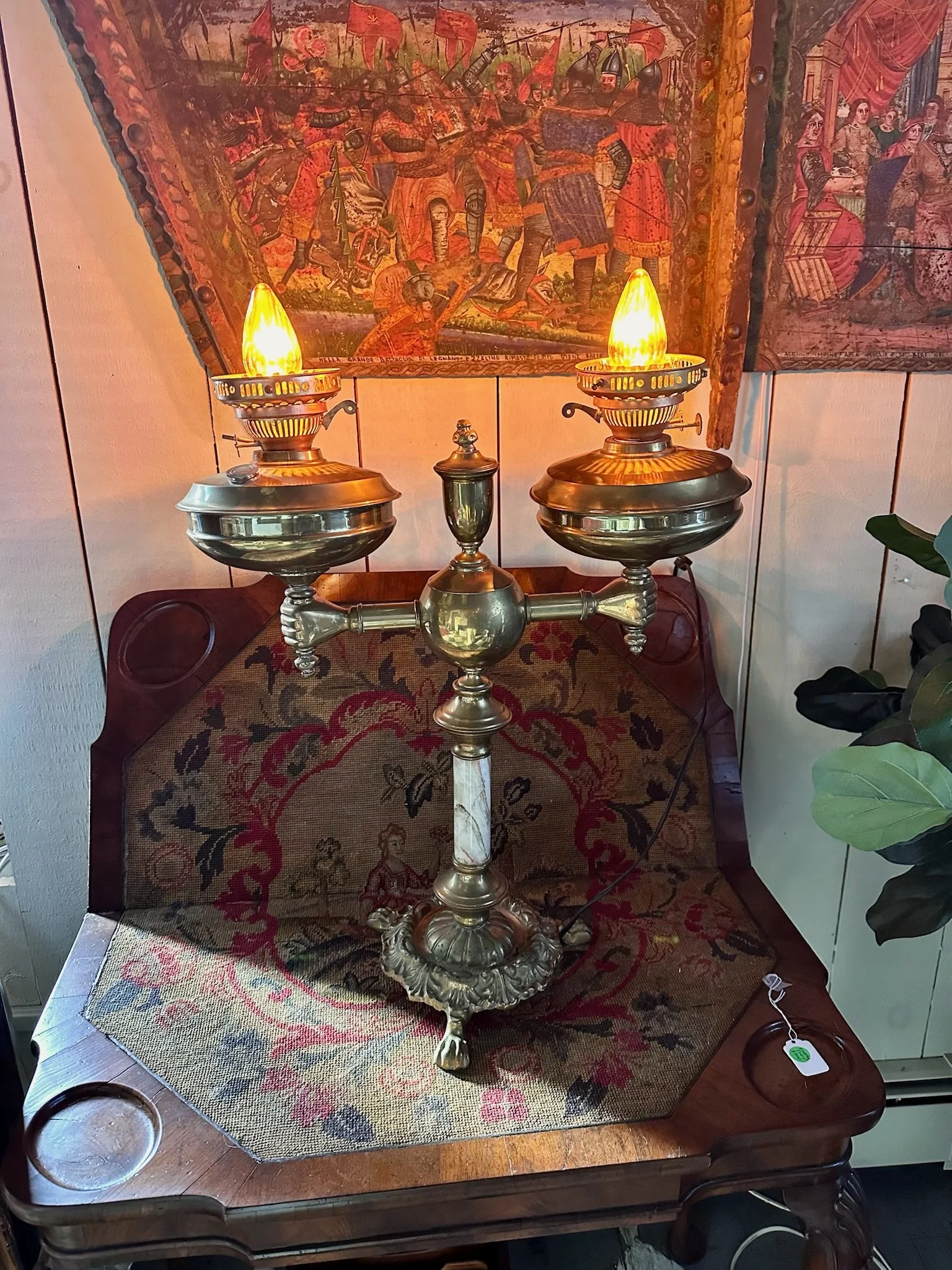 An antique oil lamp reconfigured for electricity, featuring brass hands gripping below each light. Shown on an antique needlepoint card or game table, below an antique Sicilian hand carved and hand painted donkey cart panel hanging on the wall.