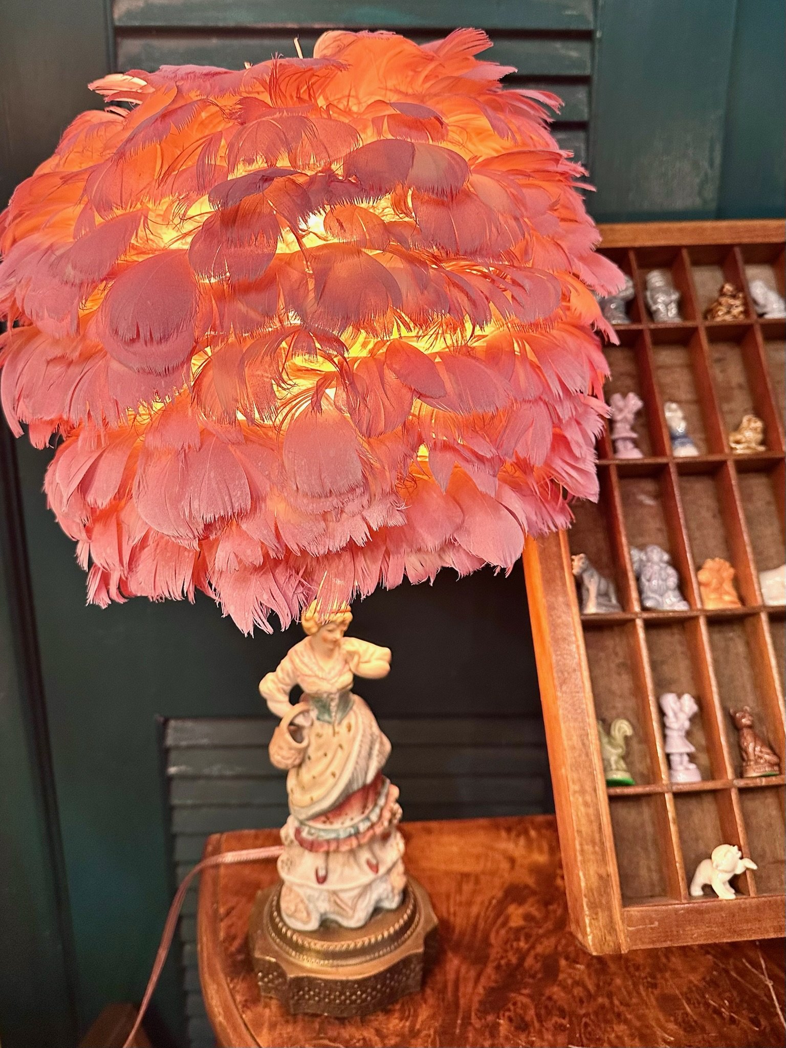 A porcelain female figural lamp with pink ostrich feather lampshade, for sale at Villa Vintiques in Woodbury, CT. In the background, a wooden shelf filled with various small figurines and sculptures, including Wade’s Whimsies from the Rose Tea boxes.